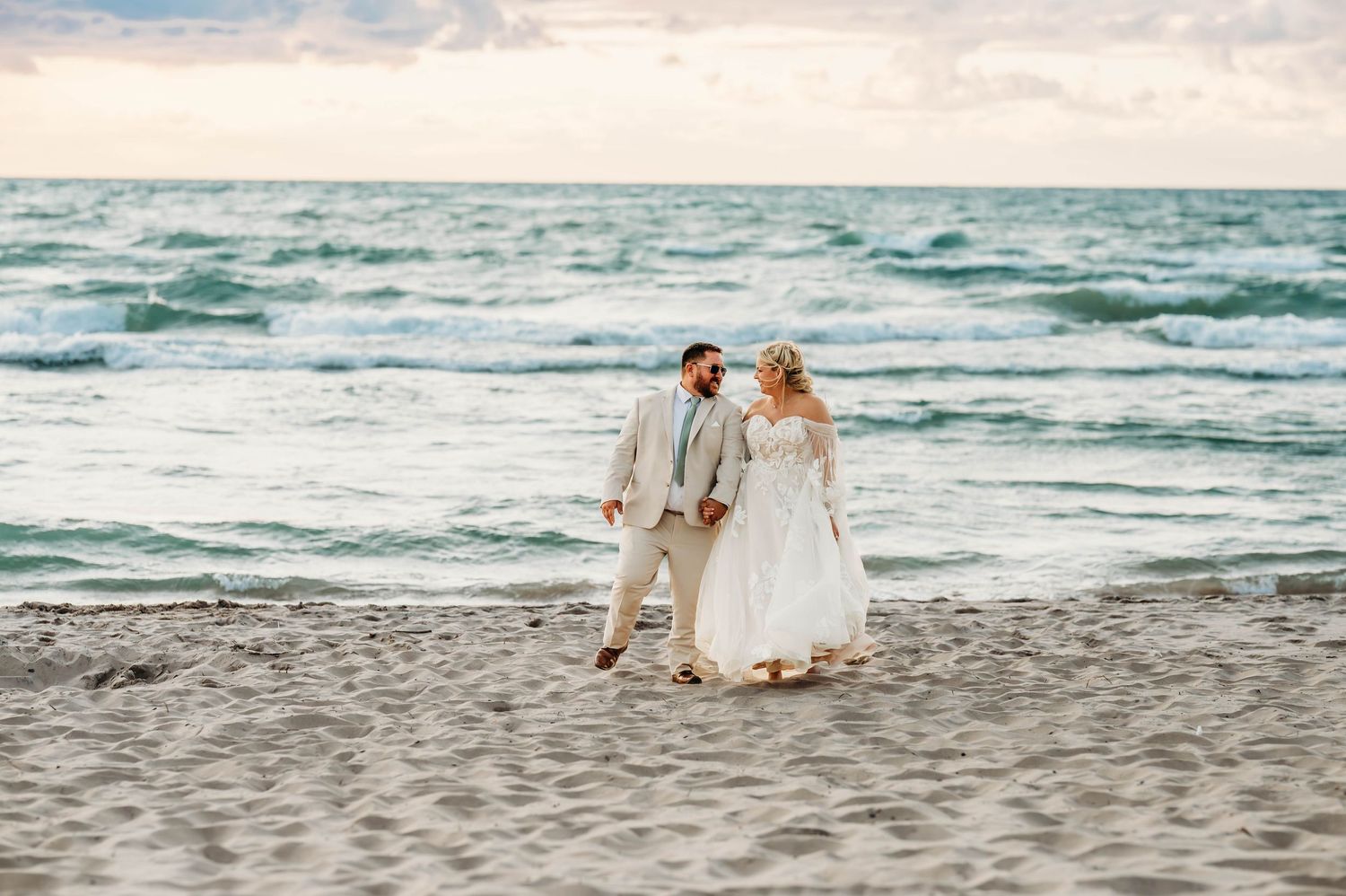 A couple in wedding attire walks along a scenic beach with waves crashing in the background at sunset.