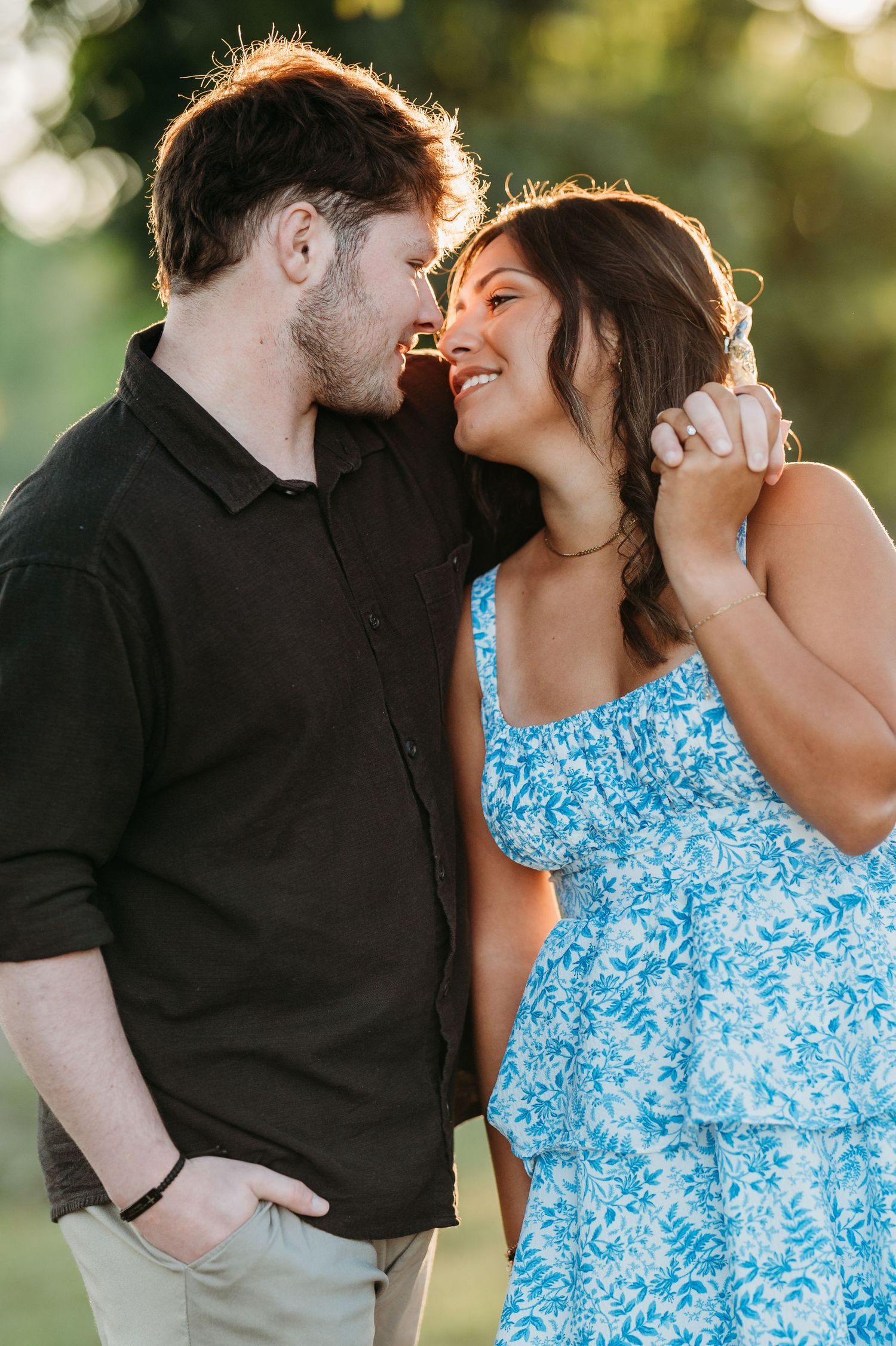 A couple in a black shirt and blue floral dress share intimate moments outdoors during a golden hour photoshoot.