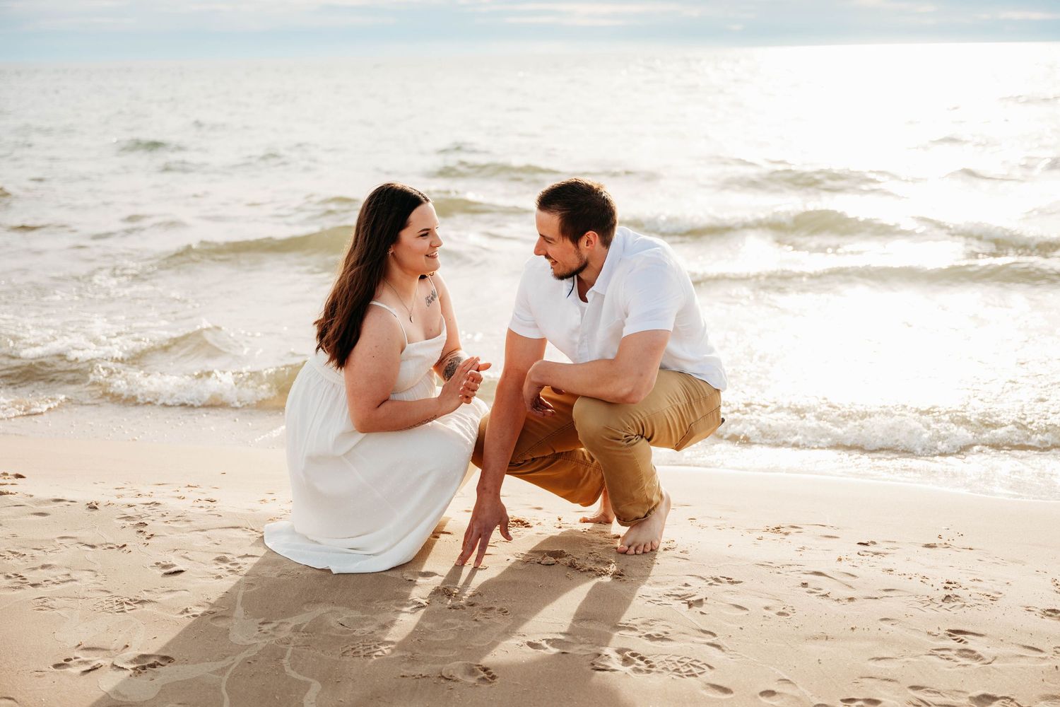 Couple sitting on sandy beach at sunset, writing in the sand near ocean waves.