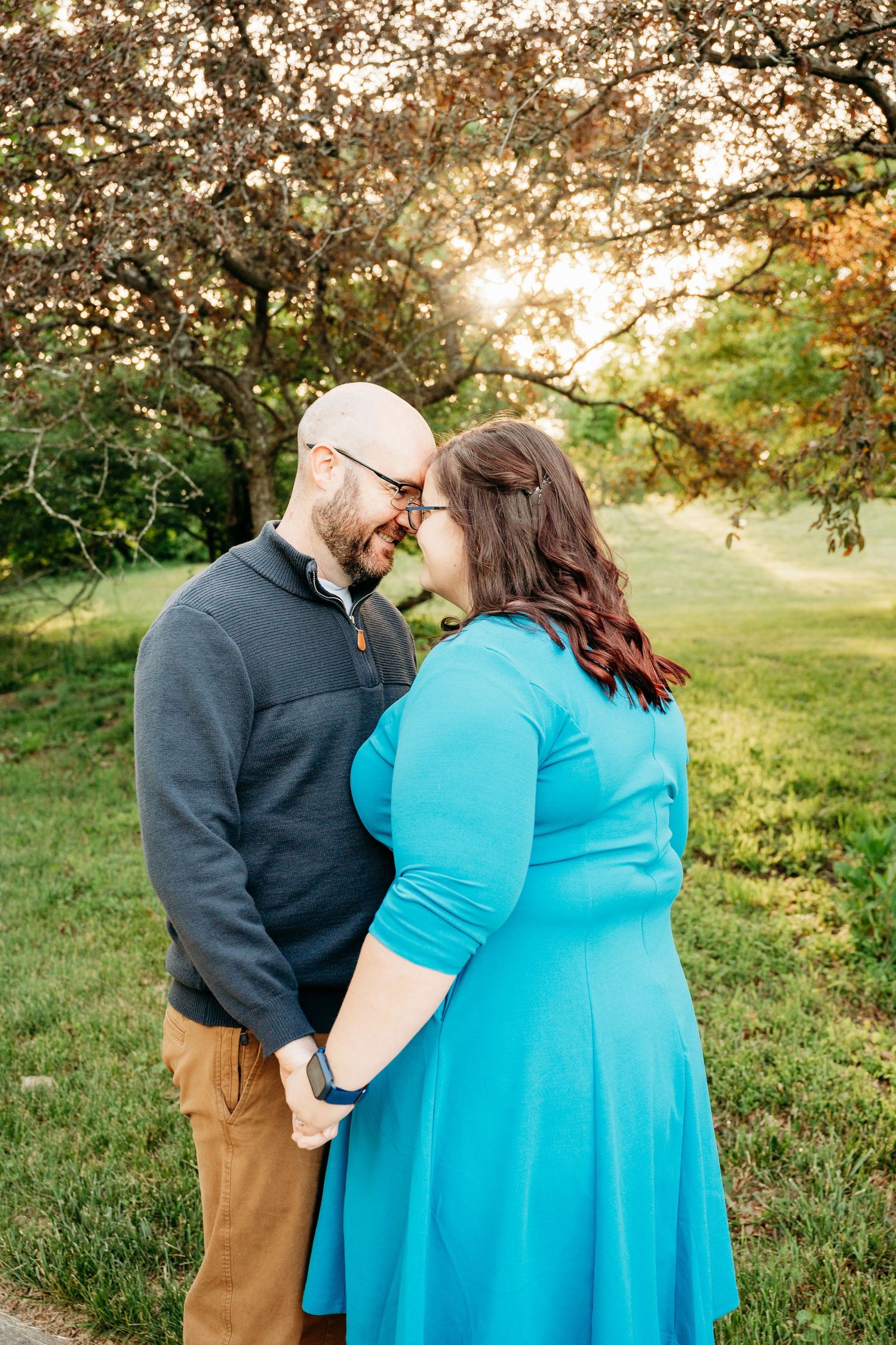 A couple shares a romantic moment holding hands in a sunlit park, the woman in a teal dress and man in a gray pullover.