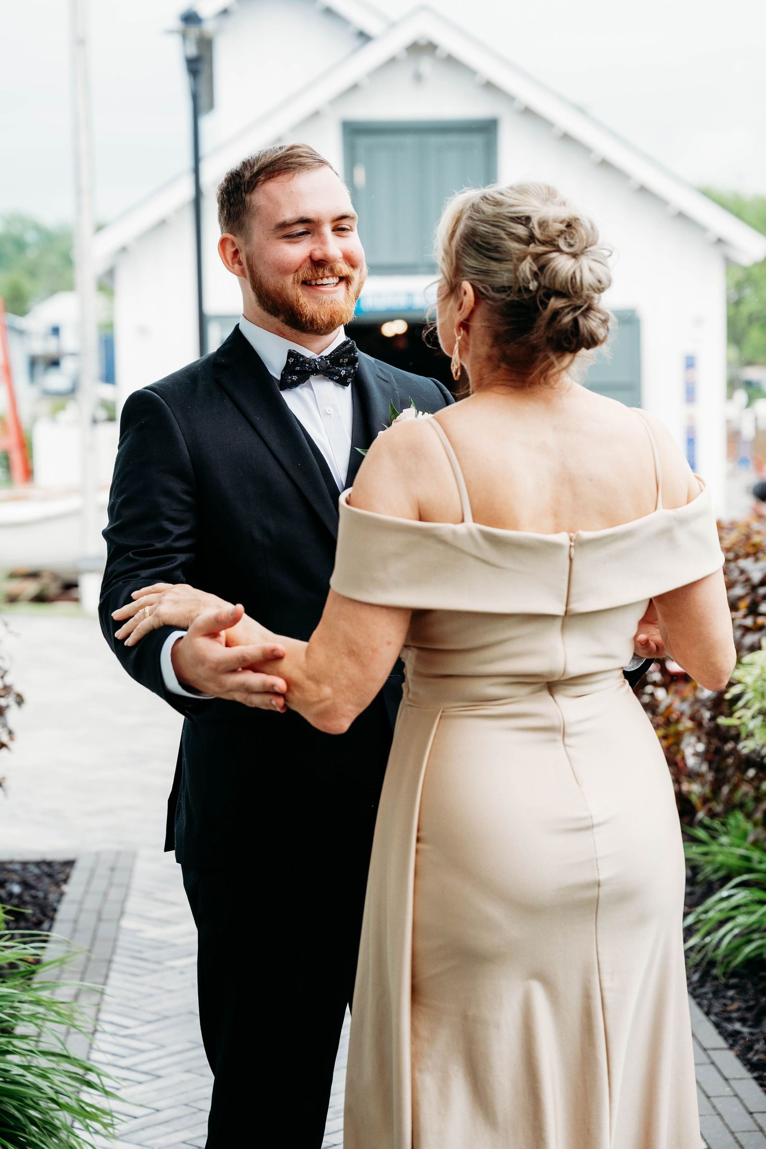 Smiling groom in black tuxedo and bow tie holds hands with woman in elegant off-shoulder champagne gown outdoors.