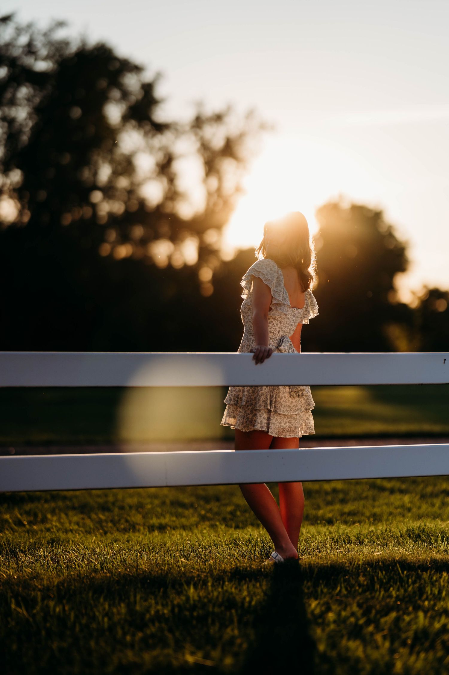 Woman in floral dress leaning on white fence bathed in warm sunset backlight on green lawn.
