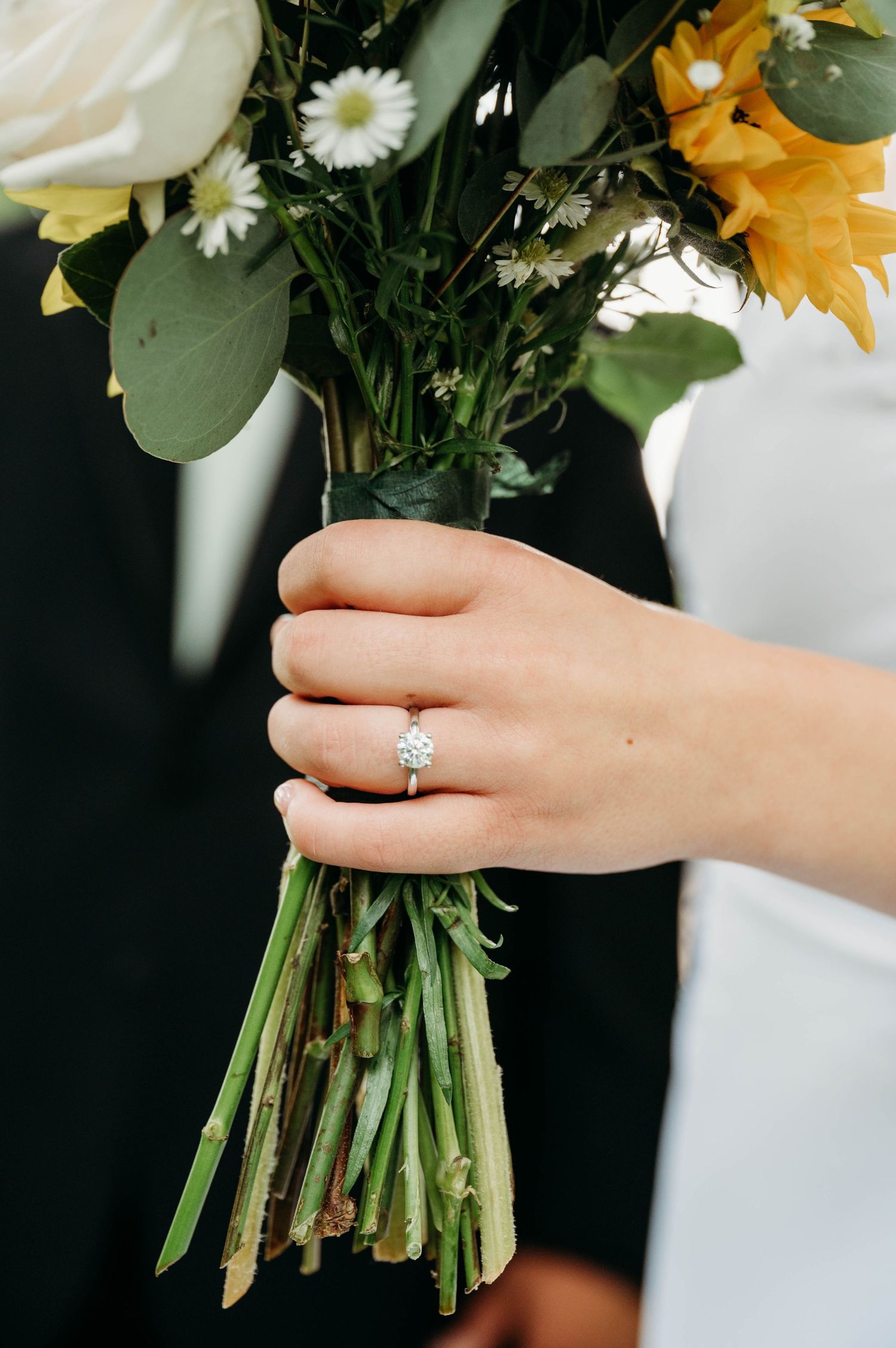 Bride holds sunflower and daisy wedding bouquet while wearing a solitaire diamond engagement ring on her finger.