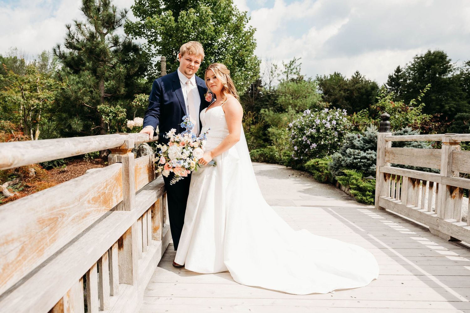 Bride and groom pose on a wooden bridge surrounded by lush greenery, bride holding a pink and white floral bouquet.