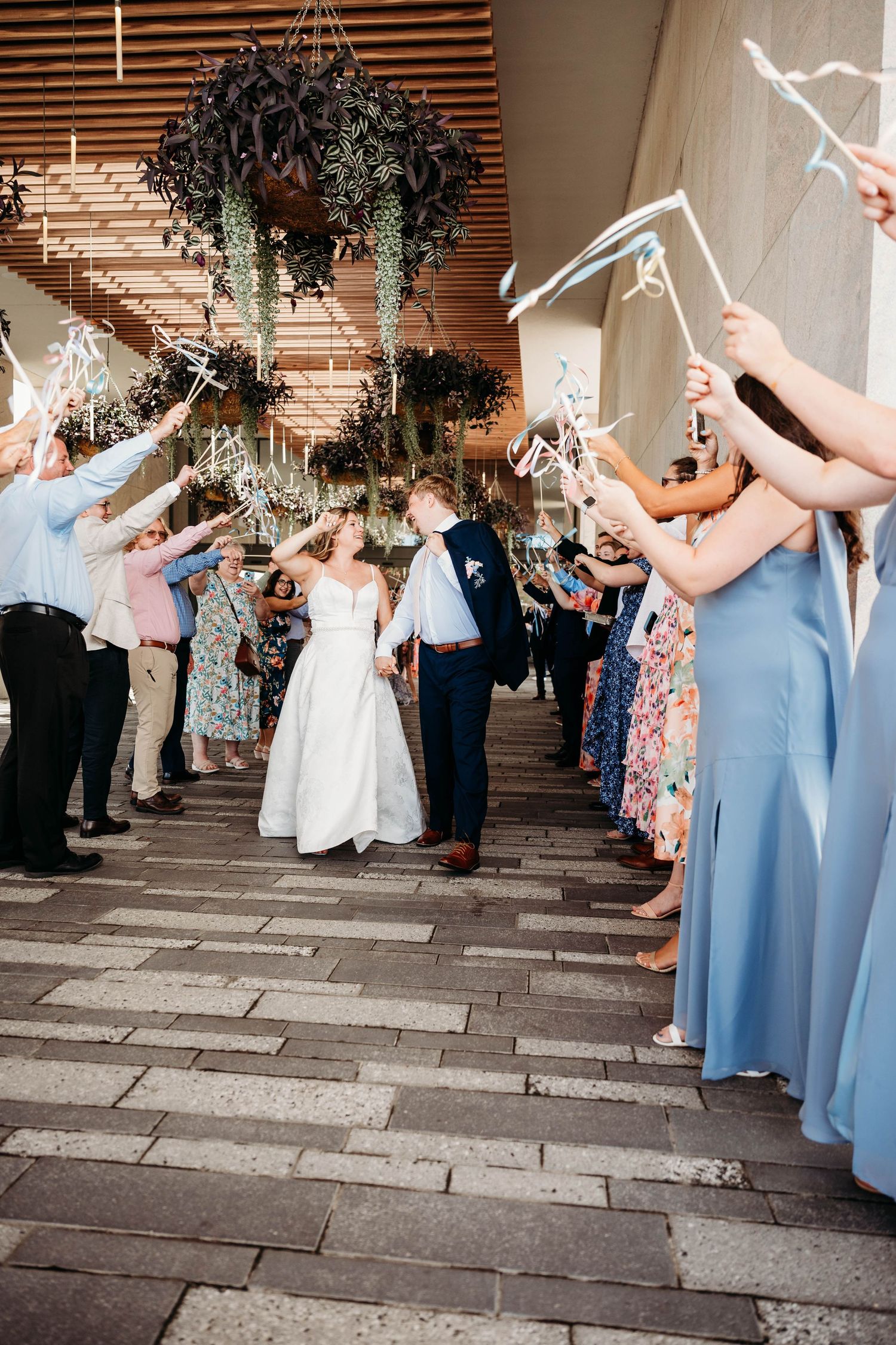 Bride and groom walk through a ribbon wand sendoff tunnel surrounded by cheering wedding guests at an elegant venue.