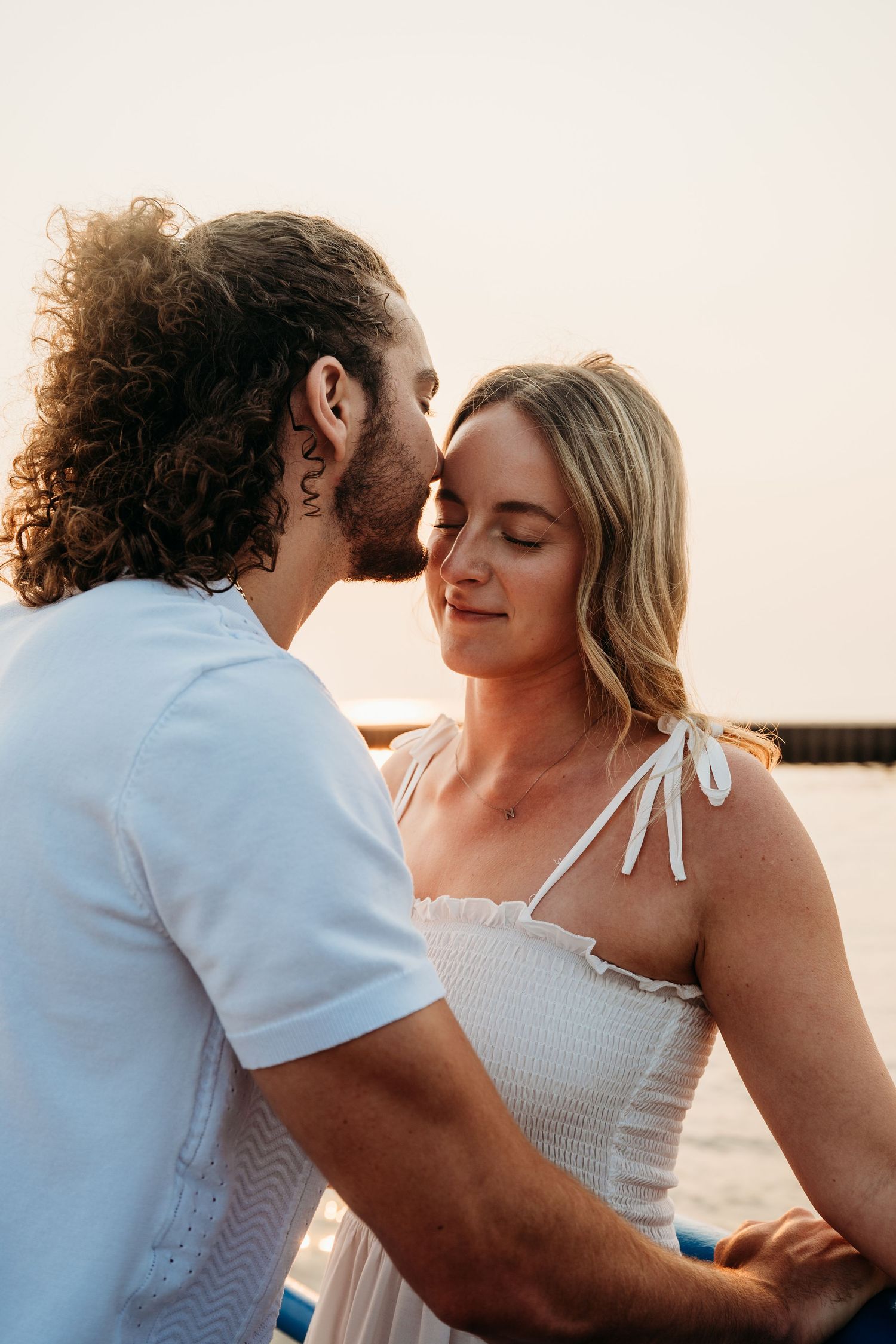 A couple shares a tender forehead kiss at sunset near the water, the woman in a white dress smiling softly.