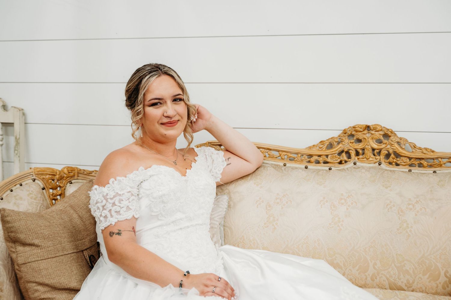 A person in an elegant off-shoulder white wedding dress sits on an ornate vintage beige sofa.