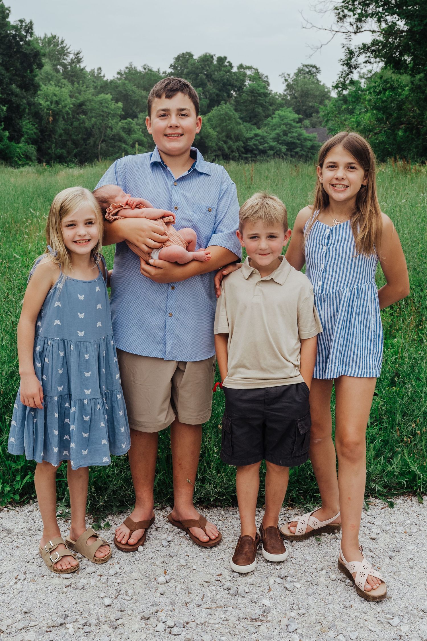 A group of children stands together on a gravel path wearing casual summer clothing in a green outdoor setting.