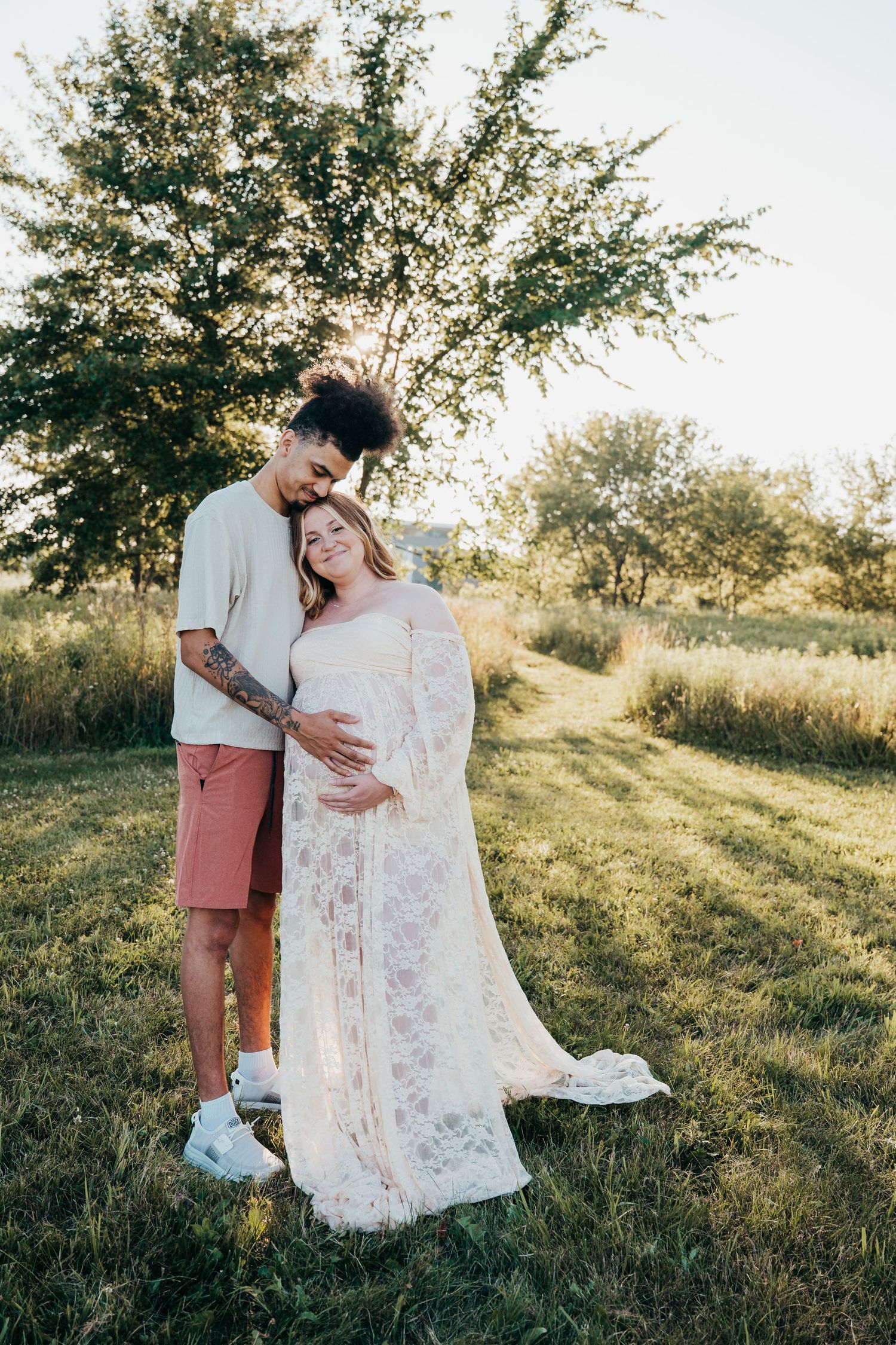 A romantic outdoor maternity photo showing a couple in a natural setting with soft sunlight filtering through trees.