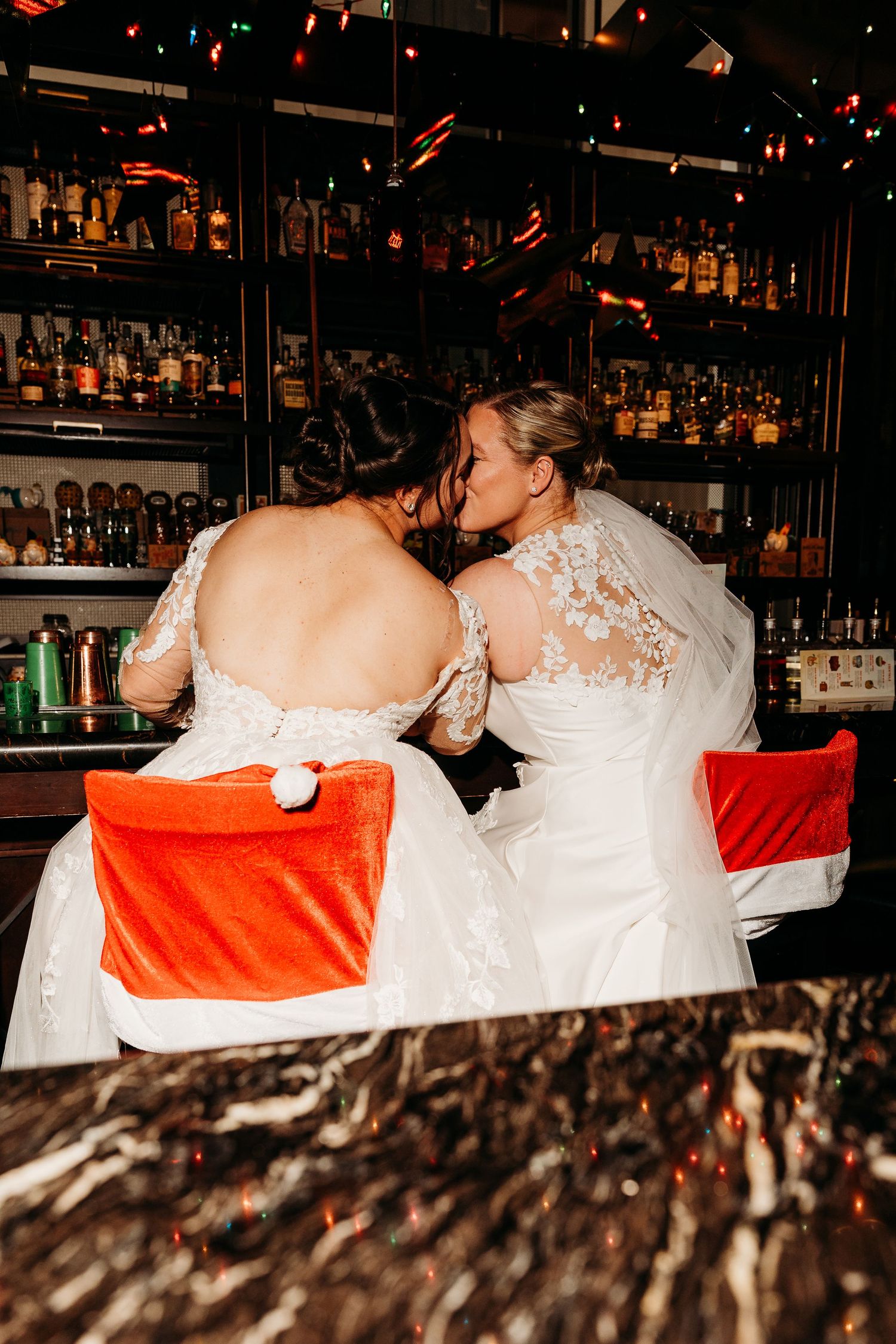 Two brides in white lace wedding dresses sit at a bar with shelves of bottles in the background.