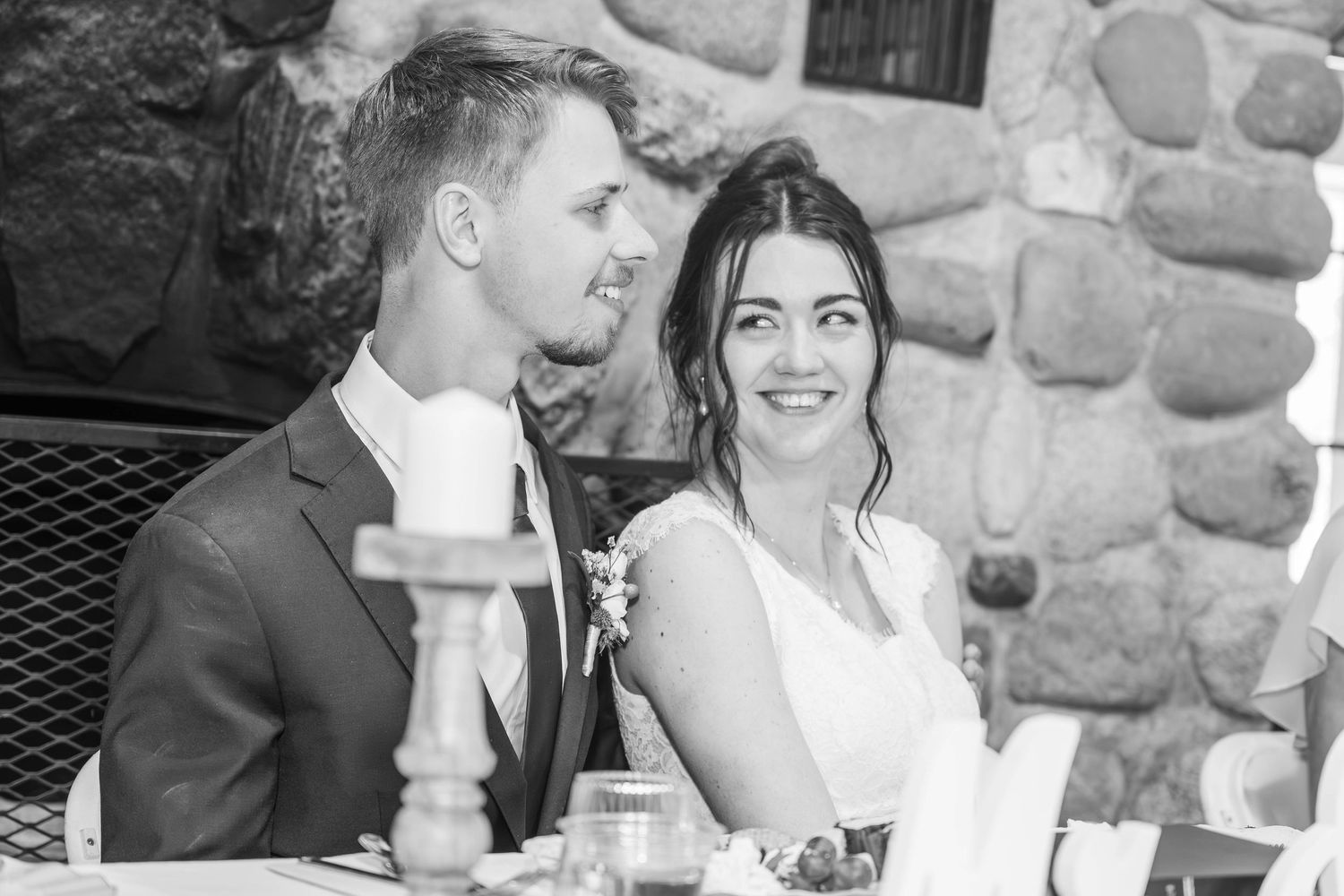 Black and white photo of a smiling bride and groom seated together at their wedding reception near a stone wall.