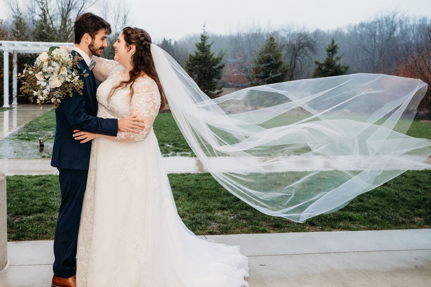 A romantic wedding moment captured as a flowing veil catches the wind on an outdoor terrace with mountain views.