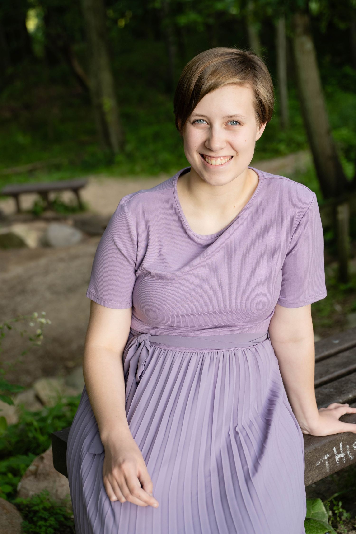 Someone wearing a lavender dress seated on a wooden bench in a forest setting.