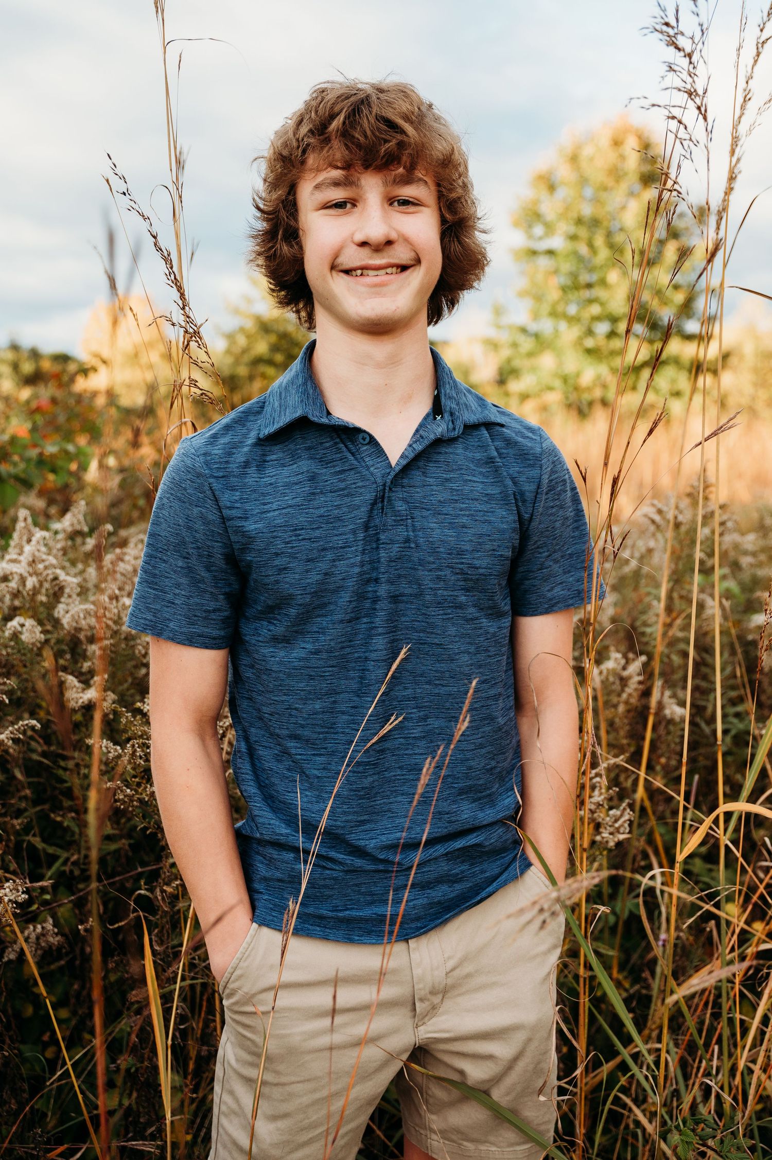 Outdoor portrait of someone in a navy blue polo shirt and khaki pants standing among tall grass during golden hour.