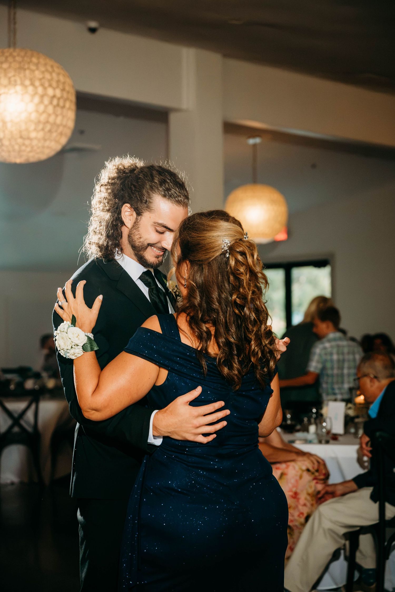 Groom in black tux shares emotional mother-son dance with woman in navy blue gown at elegant wedding reception.