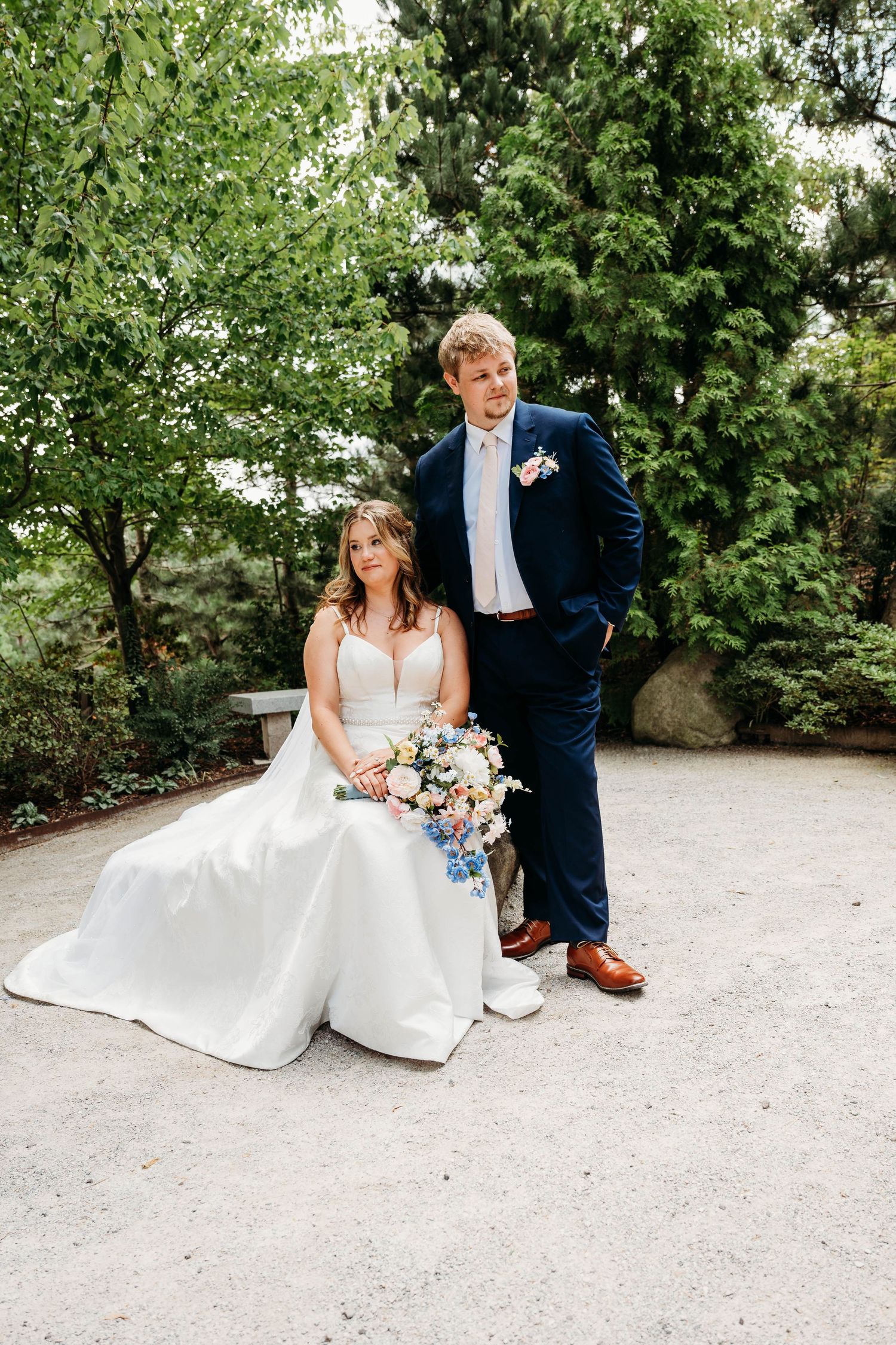 Bride in white gown sits holding floral bouquet beside groom in navy suit, posing in lush garden setting.