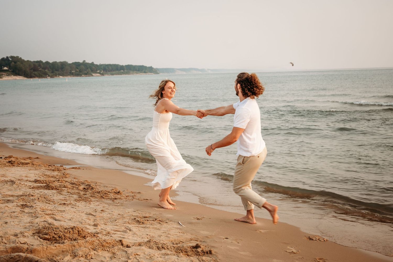 A joyful couple dances barefoot on a sandy beach at sunset, holding hands in white outfits by the ocean waves.