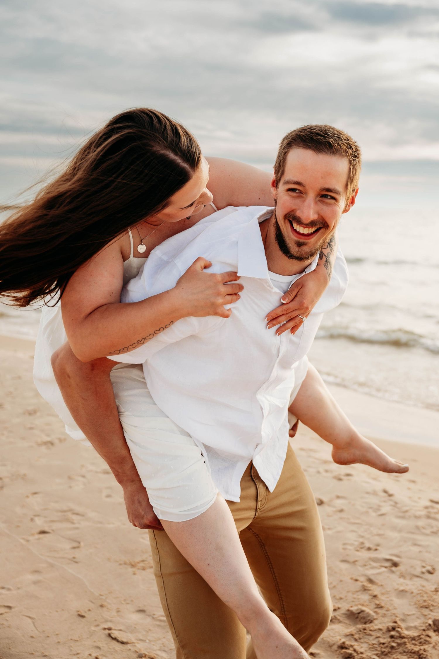 Smiling man giving woman a piggyback ride on sandy beach, both wearing white tops and tan shorts.