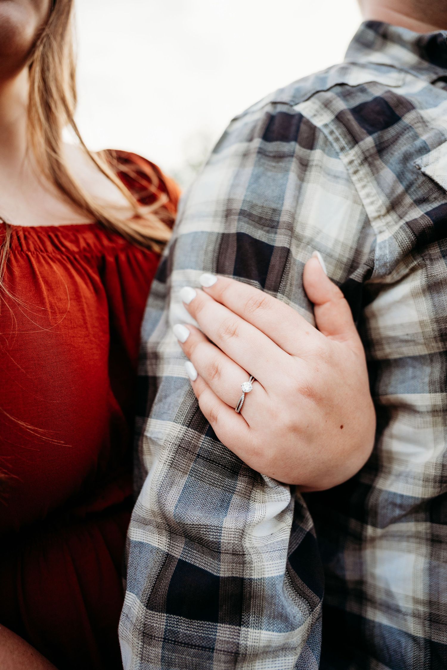 Close-up of a woman wearing an engagement ring, holding a man's arm in a plaid shirt, with her red dress visible.