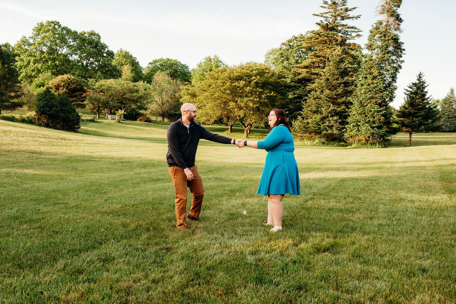 Couple standing on lush green lawn surrounded by tall trees during golden hour in a park setting.