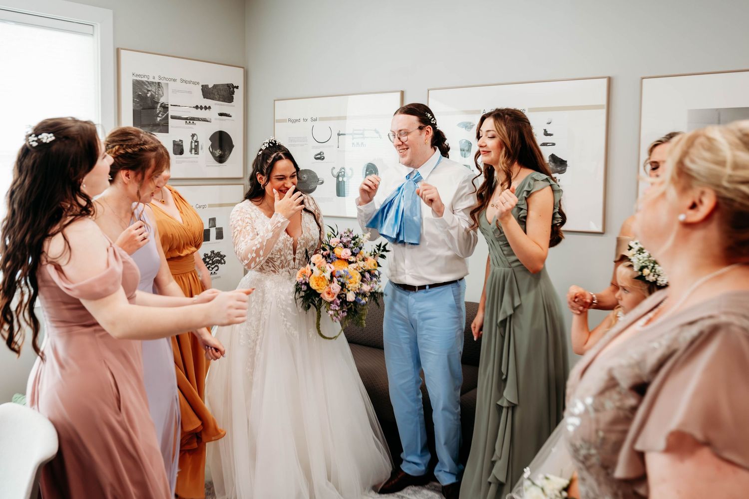 Bride and bridesmaids laughing together in dressing room, wearing blush, sage, and blue gowns with colorful bouquets.