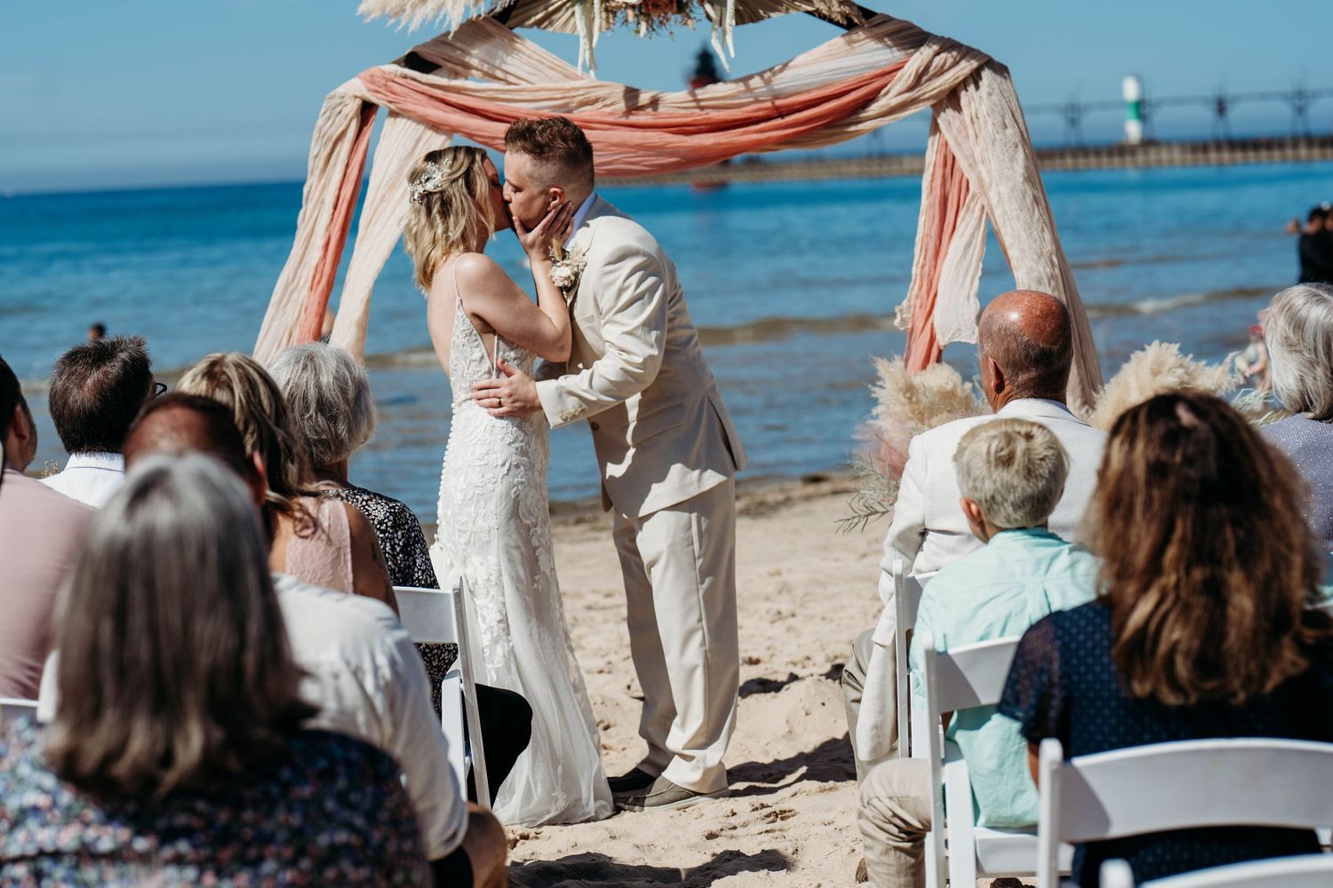 A beach wedding ceremony under a draped arch with guests seated on white chairs by the ocean.