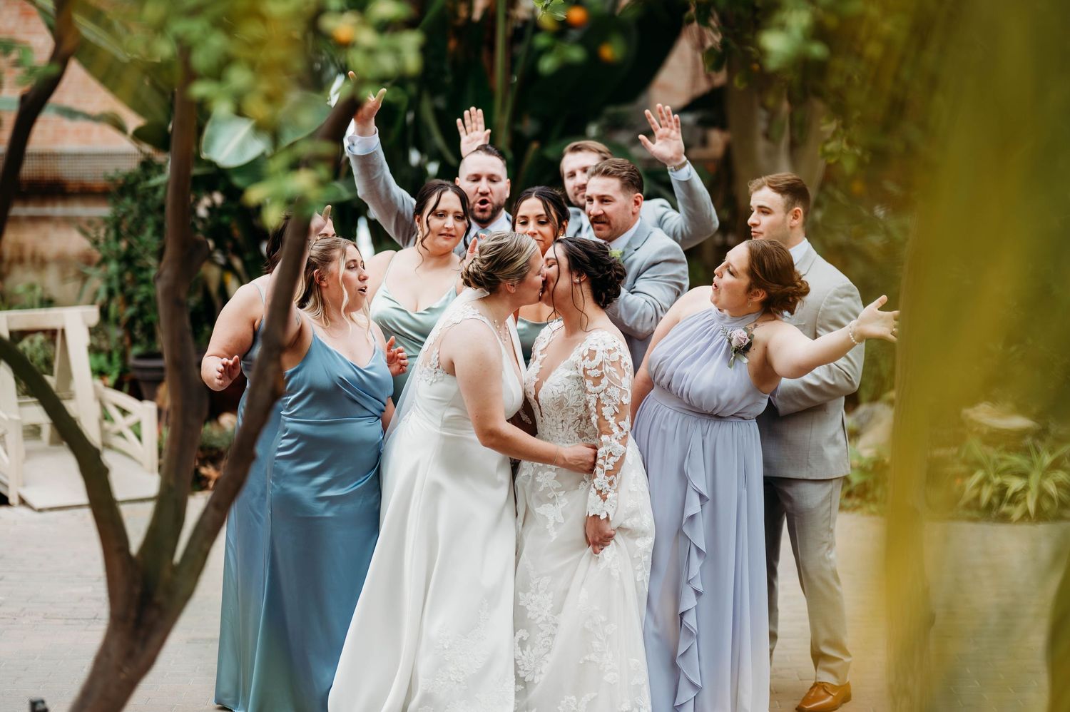 Wedding party celebrates joyfully in a garden setting wearing pastel blue dresses and light colored suits.