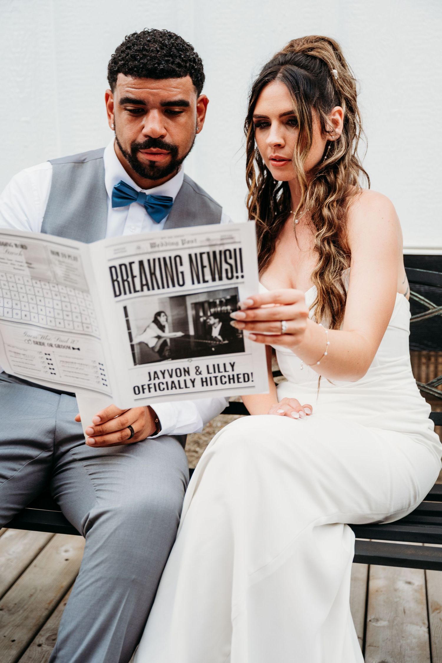 Newlyweds in wedding attire reading a newspaper with 'Breaking News' headline while sitting together on a bench.
