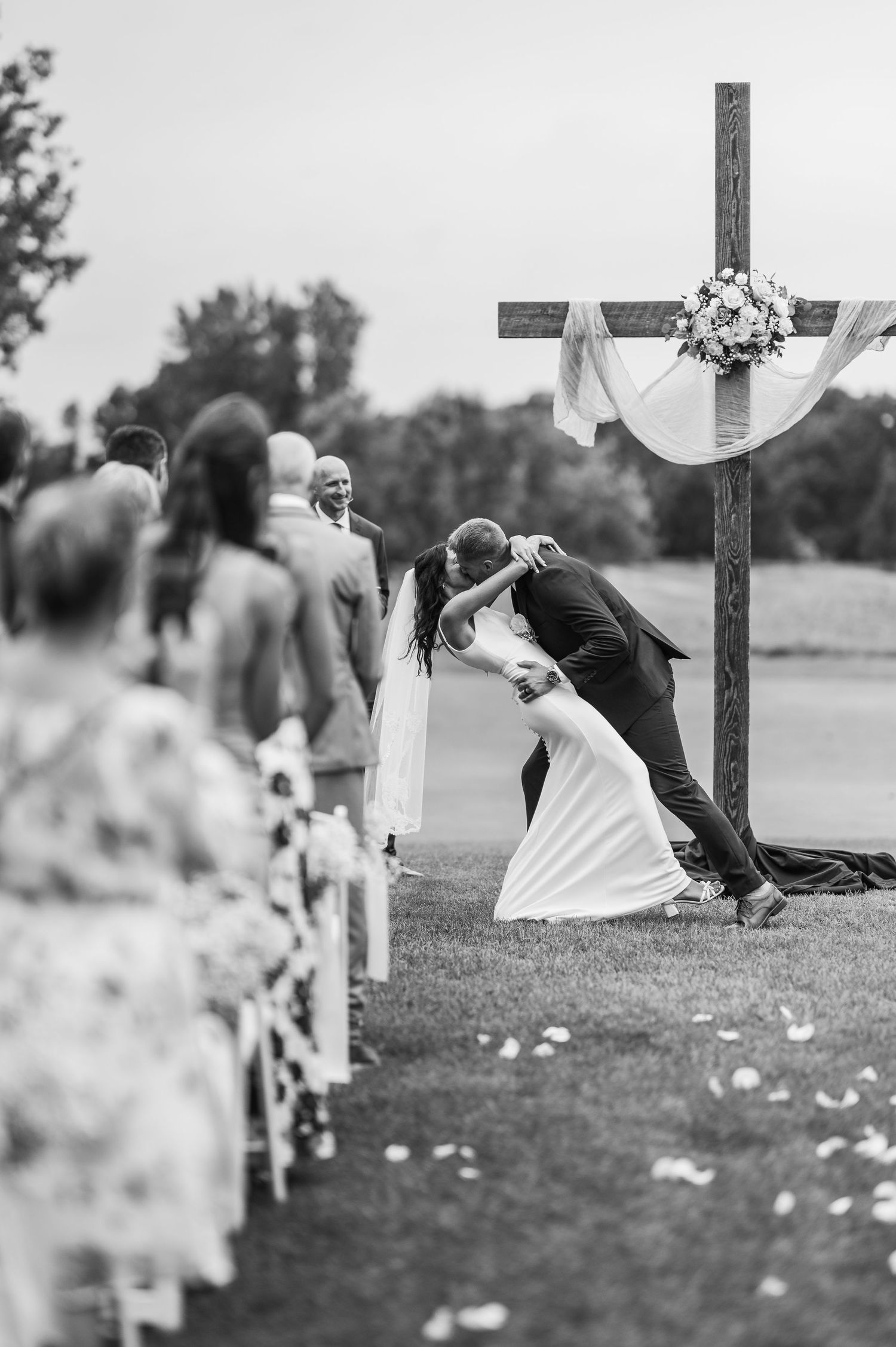 Black and white wedding ceremony photo showing a couple sharing a kiss by a wooden cross outdoors.