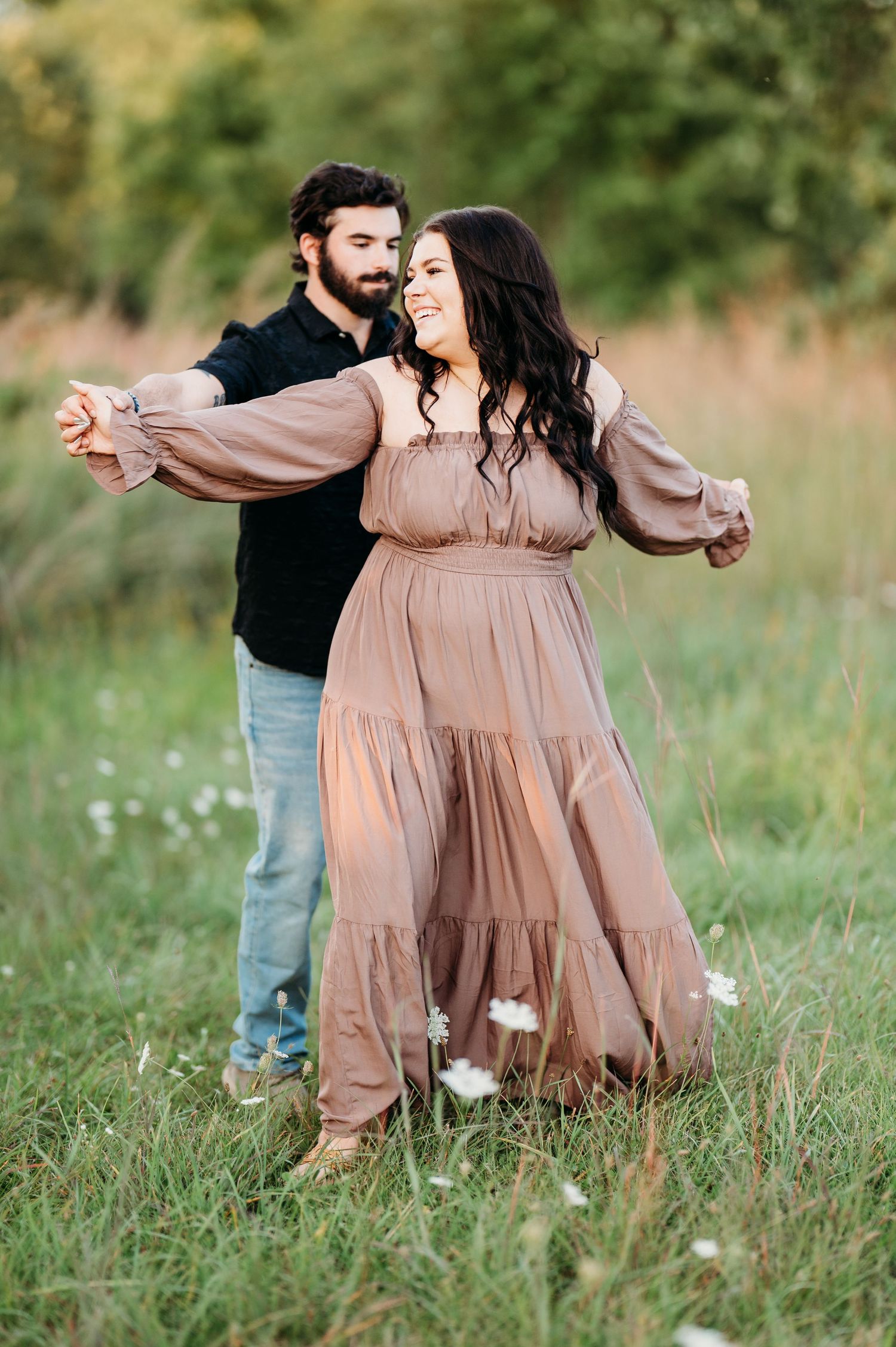 A couple embraces in a sunlit meadow with tall grass, the woman wearing a flowing beige dress.