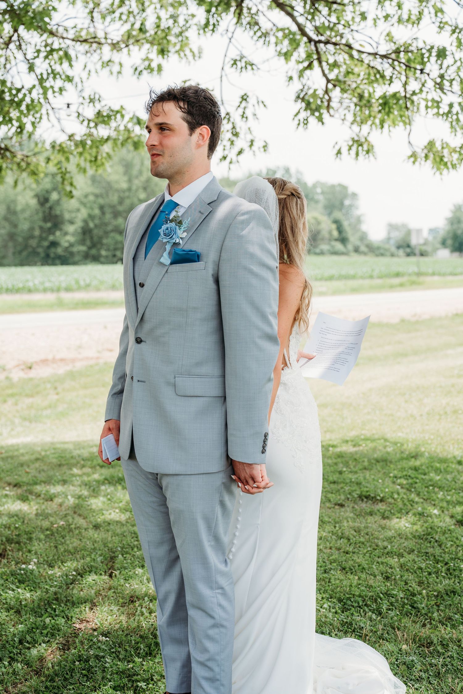 A couple in wedding attire stands together under a willow tree in a serene outdoor setting.