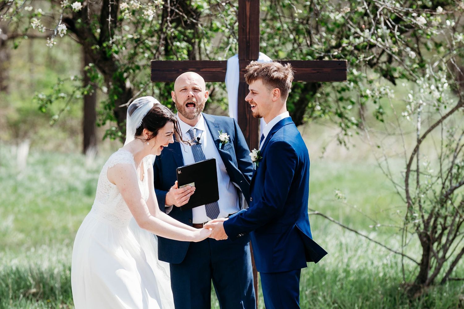 Outdoor wedding ceremony with bride and groom exchanging vows before an officiant at a wooden cross altar.
