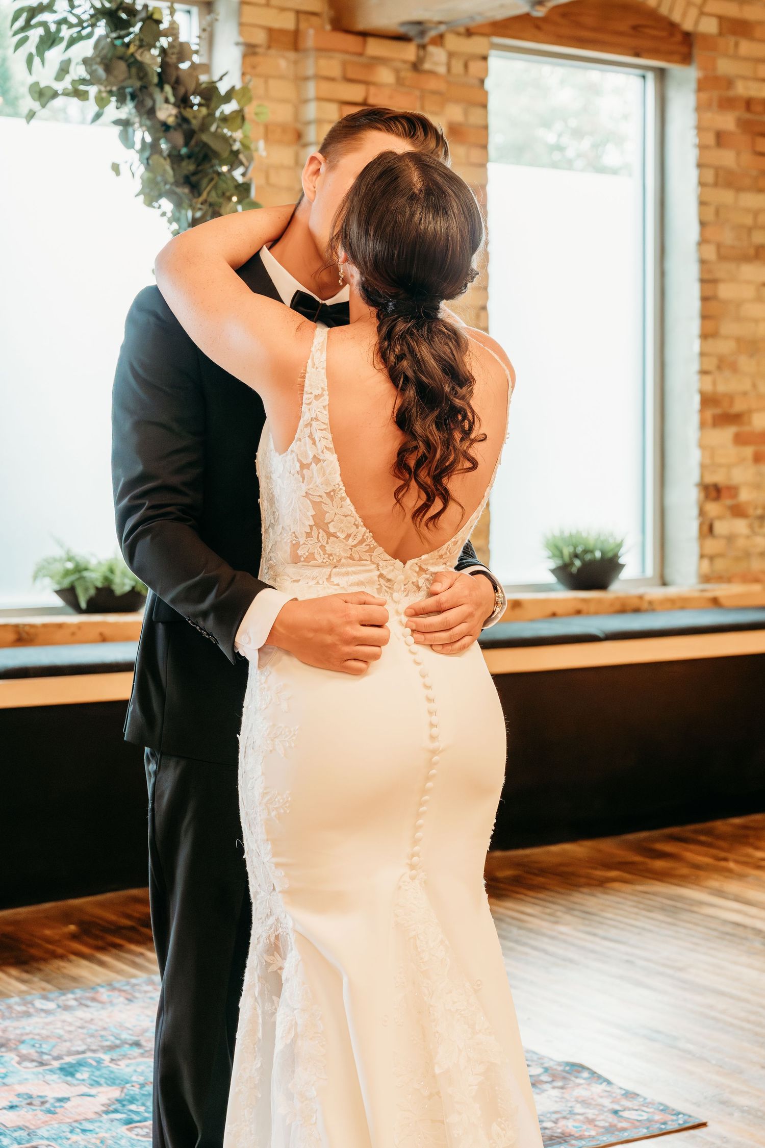 Bride and groom share first dance kiss, bride in backless lace gown with button detail in rustic brick venue.