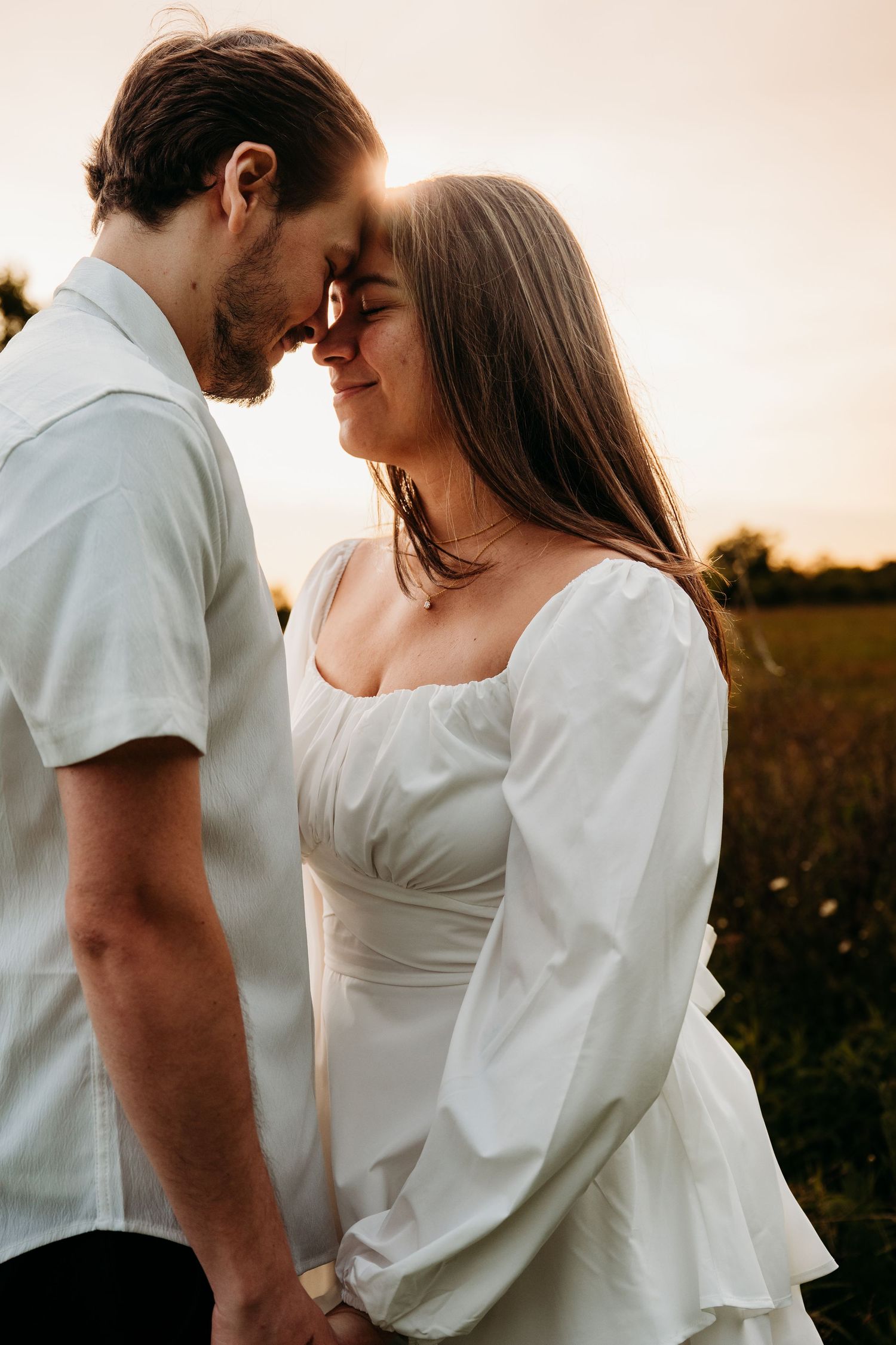 A romantic moment between a couple in a sunlit field wearing white outfits during sunset.