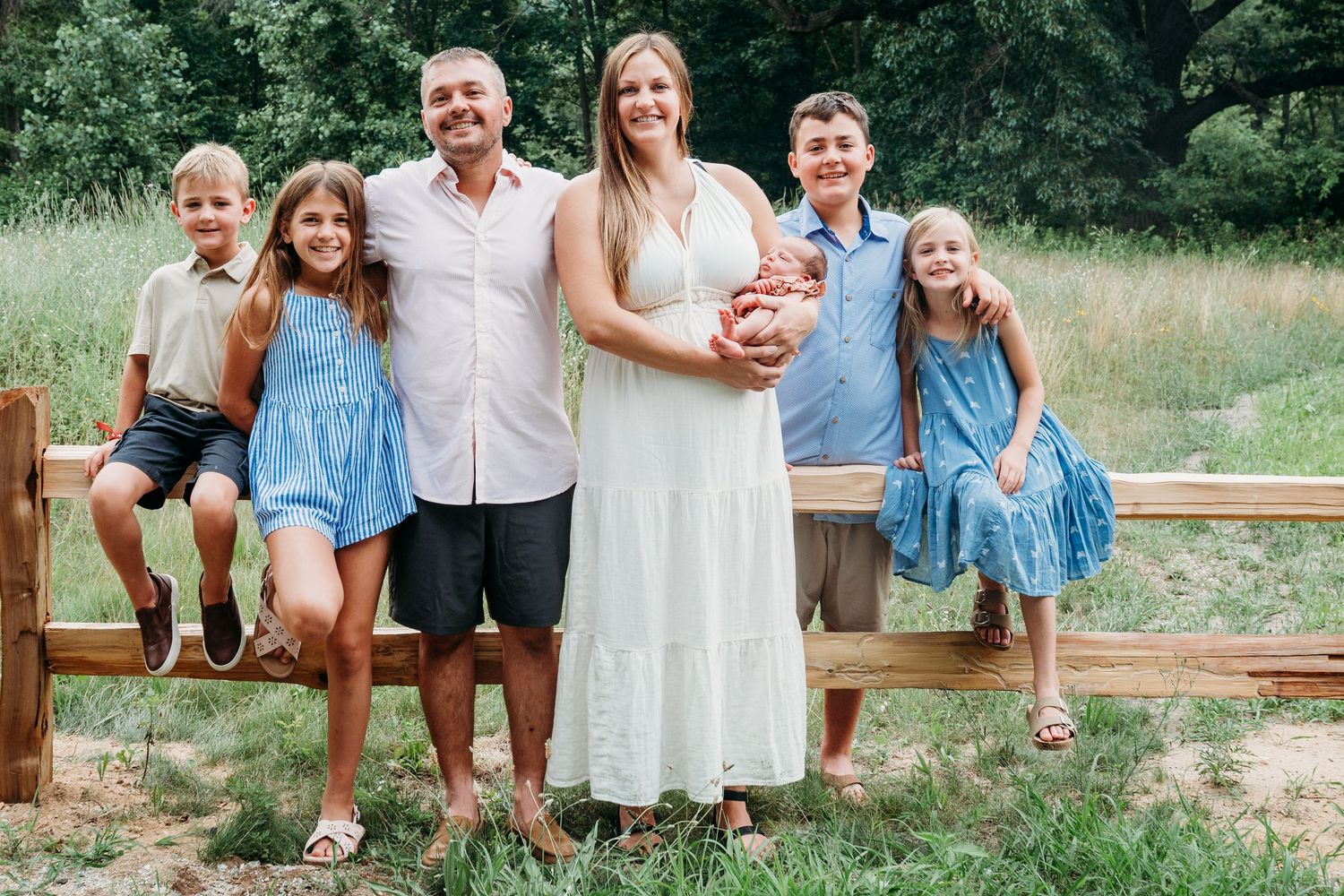 Family wearing coordinated blue and white outfits poses together on a wooden fence in a grassy outdoor setting.