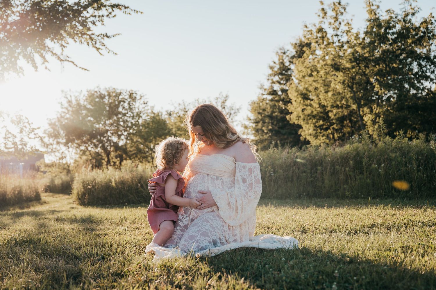 Two figures sit together on grassy hill at sunset with sunlight streaming through trees in background.