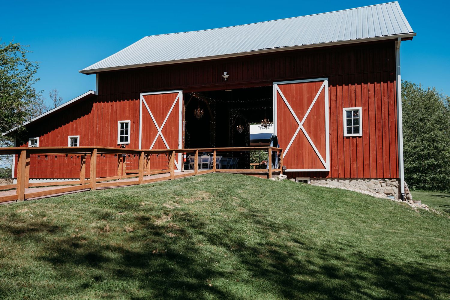 Classic red barn with white X-frame doors and metal roof stands on grassy slope on sunny day with blue sky.