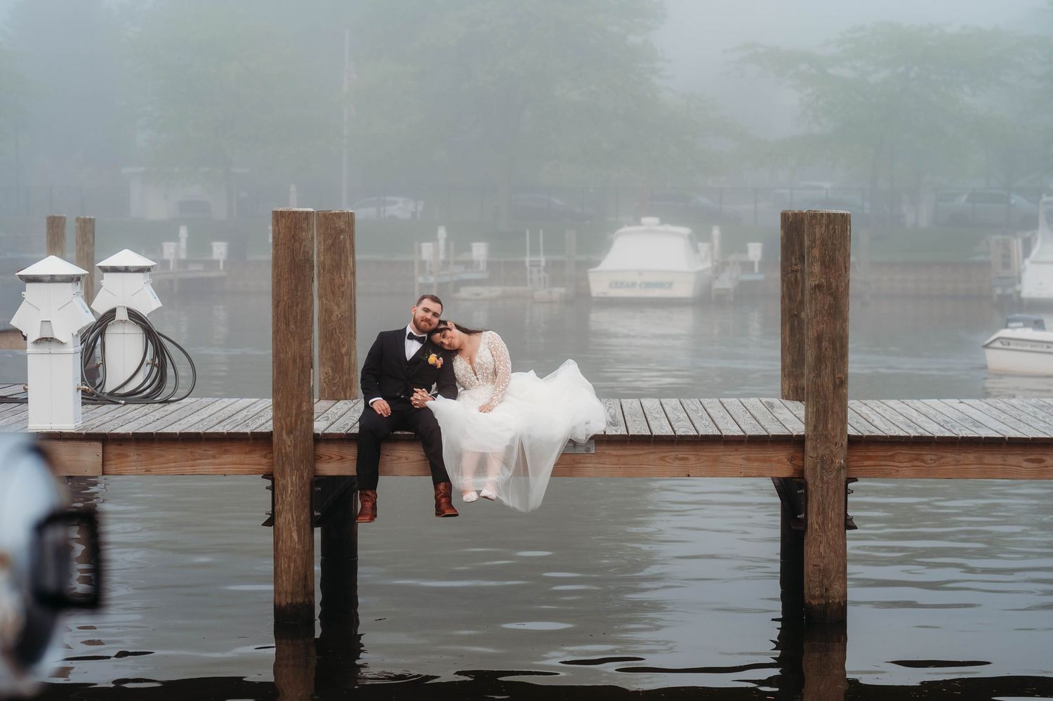Bride and groom sitting on a wooden dock over misty water, sharing a romantic kiss on their wedding day.