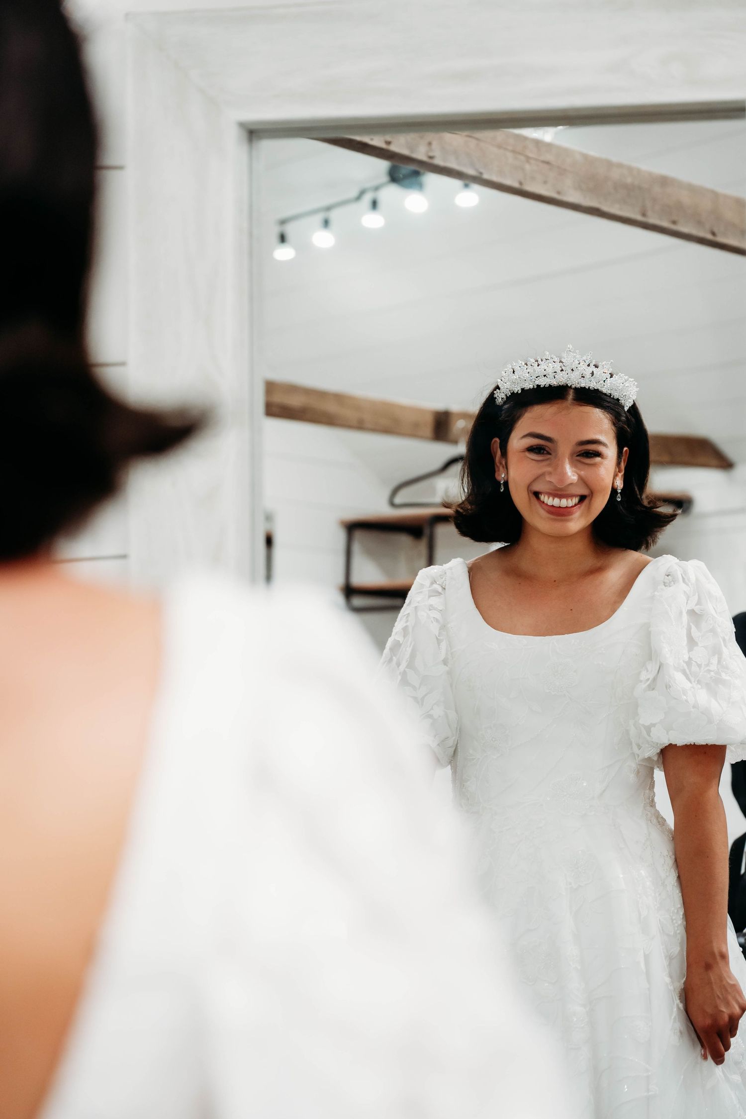 Smiling bride wearing tiara and white lace wedding dress admiring herself in mirror.