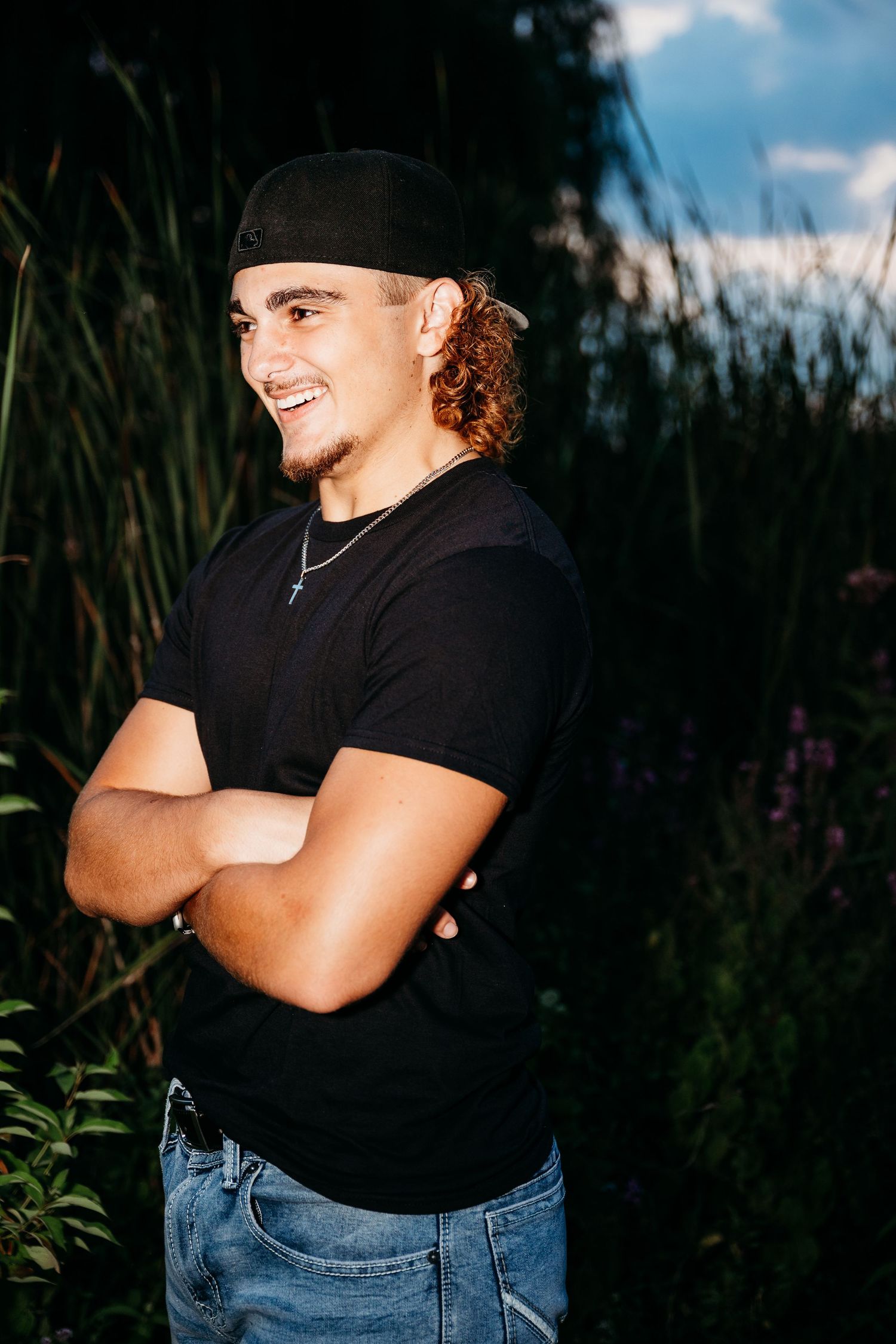 Person wearing a black t-shirt and jeans stands against dark foliage in evening light.