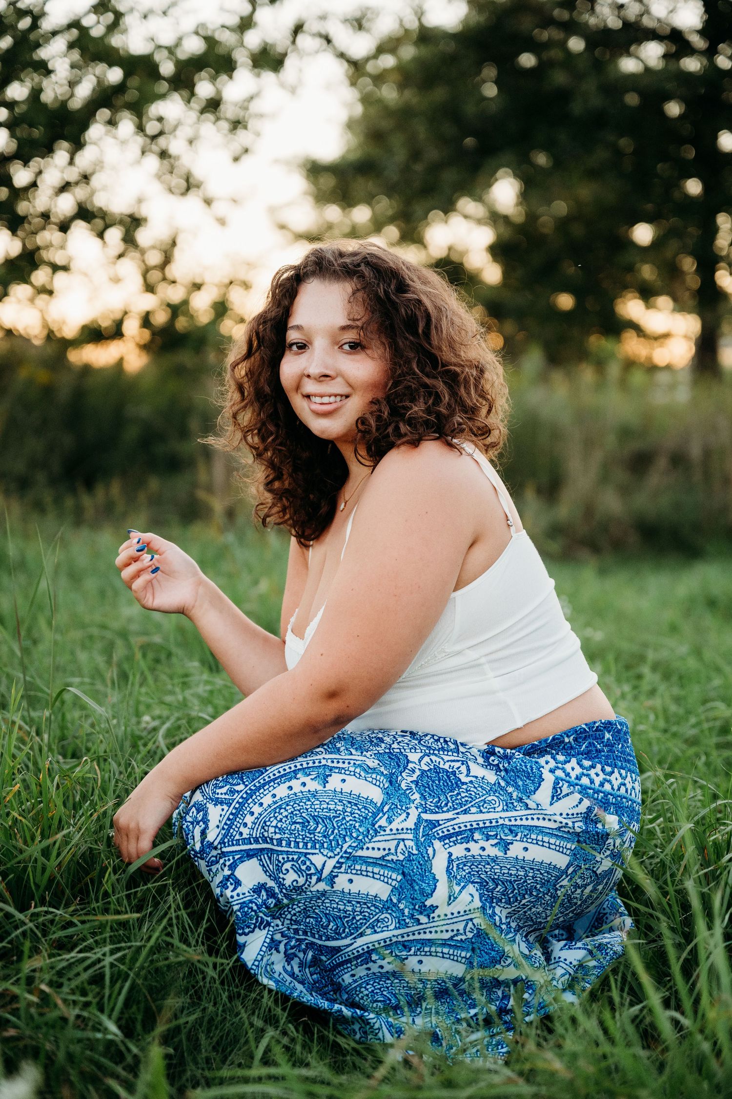 Candid lifestyle portraits showing someone in a white top and blue patterned skirt sitting in a sunlit grassy meadow.