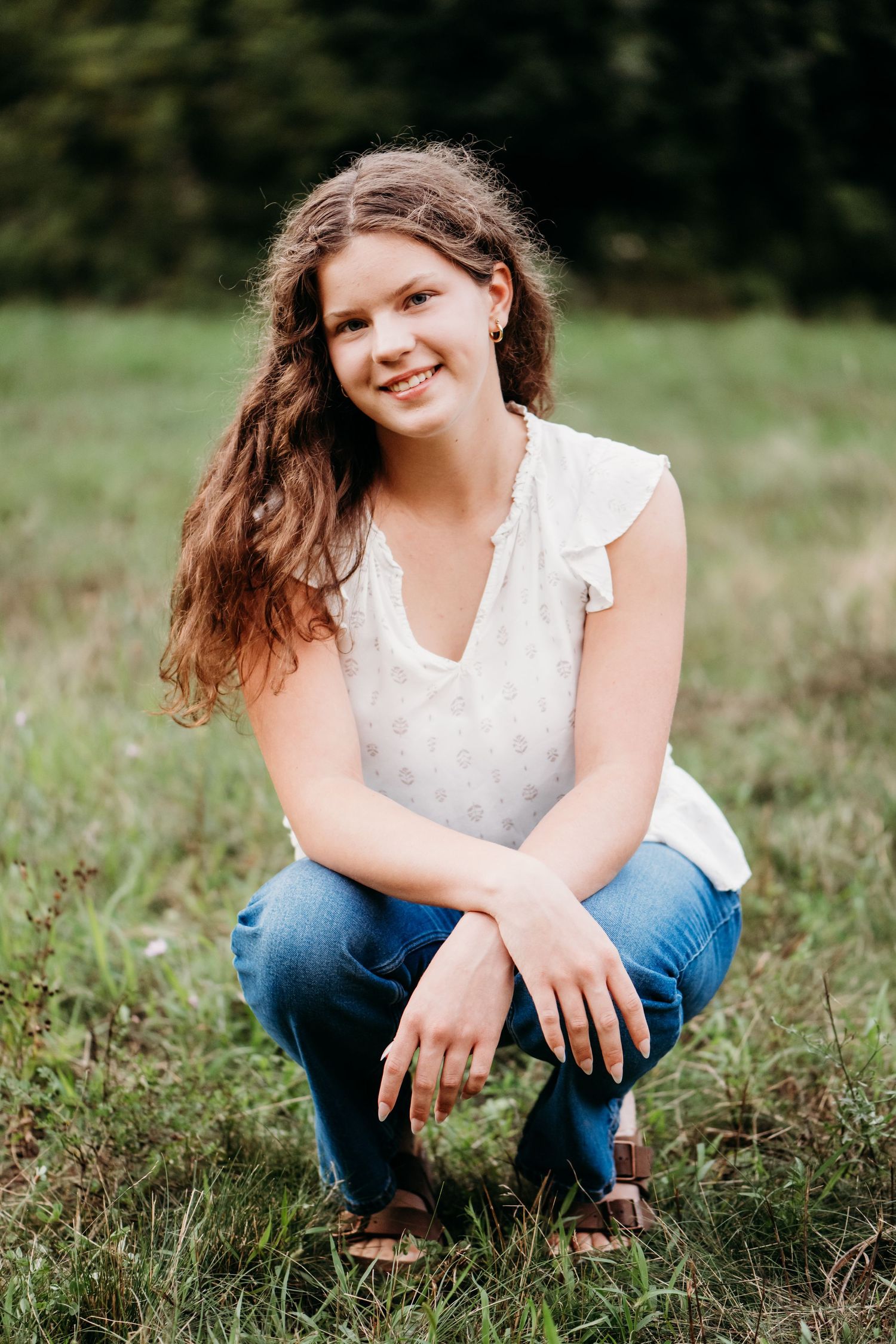 A person wearing a white lace top and blue jeans crouches in a grassy field on a sunny day.
