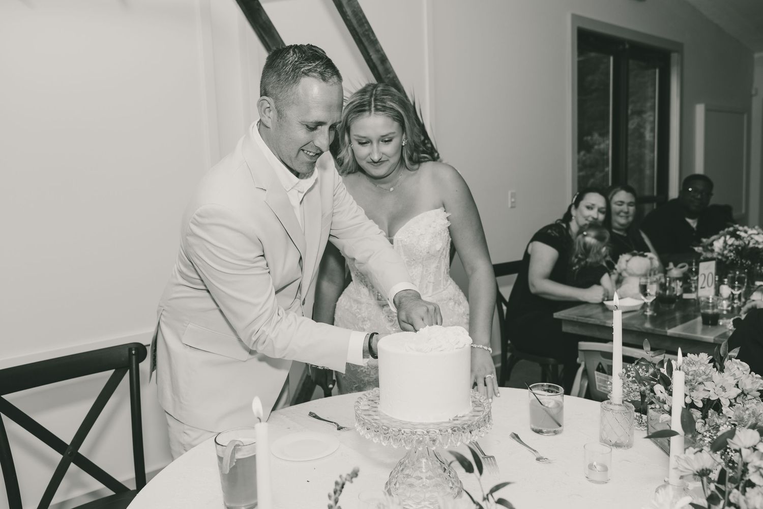 A couple cuts into their elegant white wedding cake at a reception while guests watch from tables in the background.