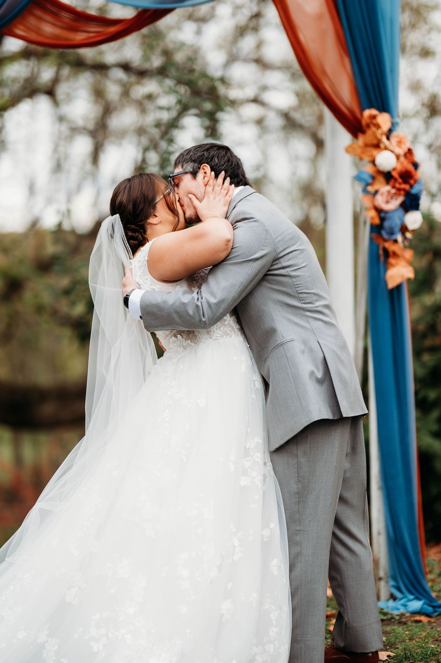A romantic wedding ceremony kiss under a blue and orange draped arch in an outdoor garden setting.