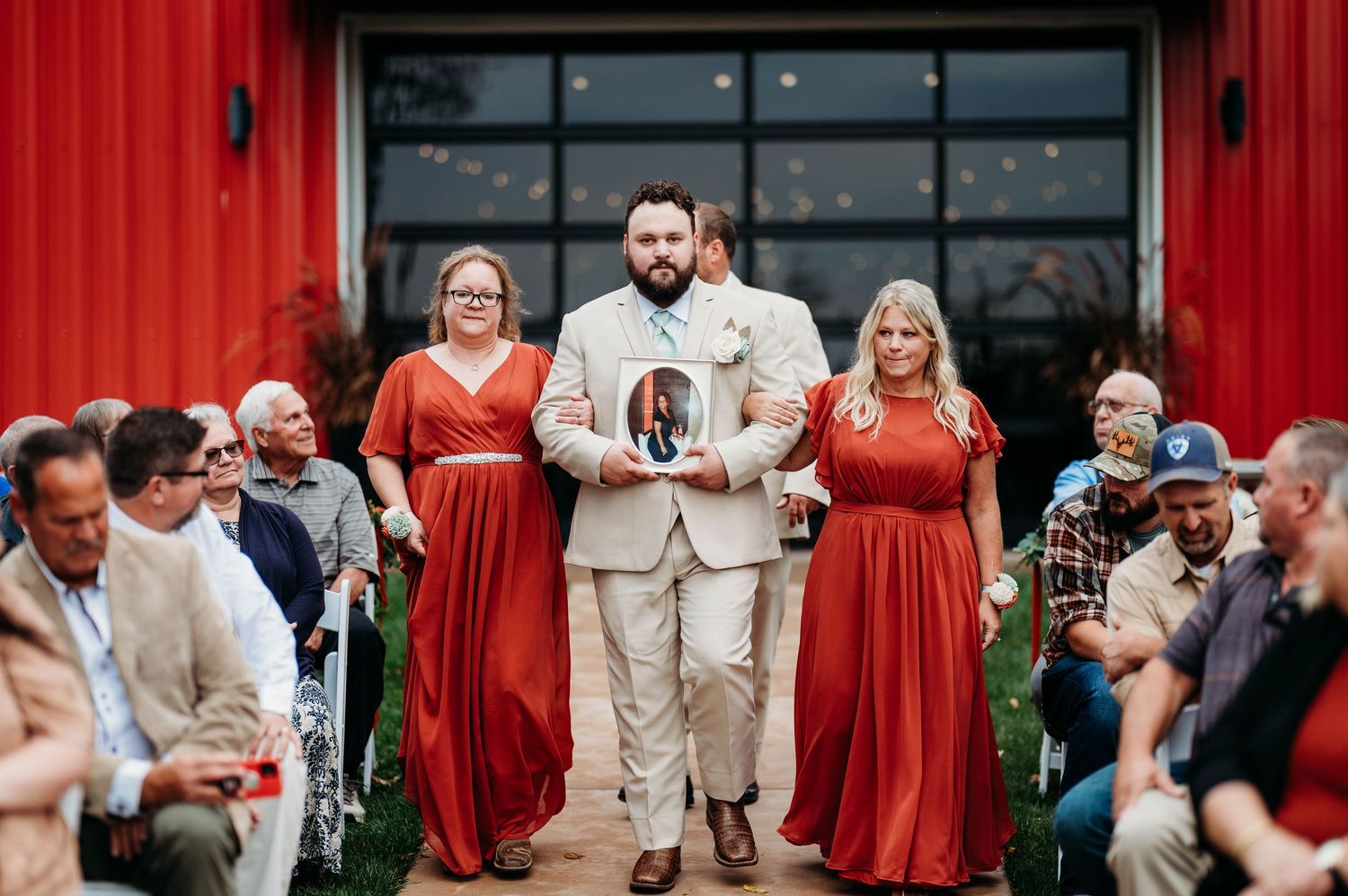 Wedding party in red dresses and beige suits poses outdoors in front of glass windows.