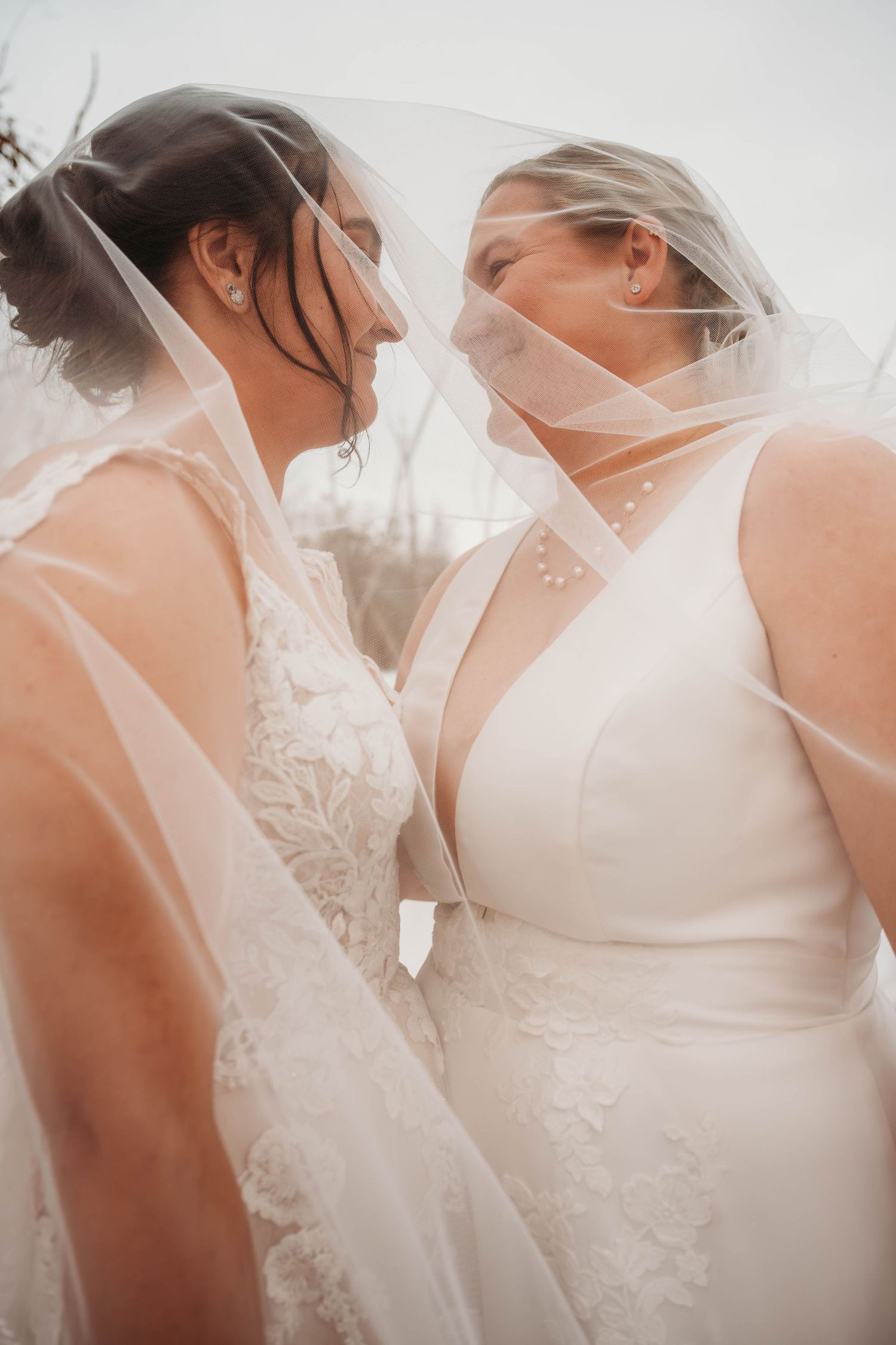 Two brides in elegant white gowns share an intimate moment under their wedding veils.