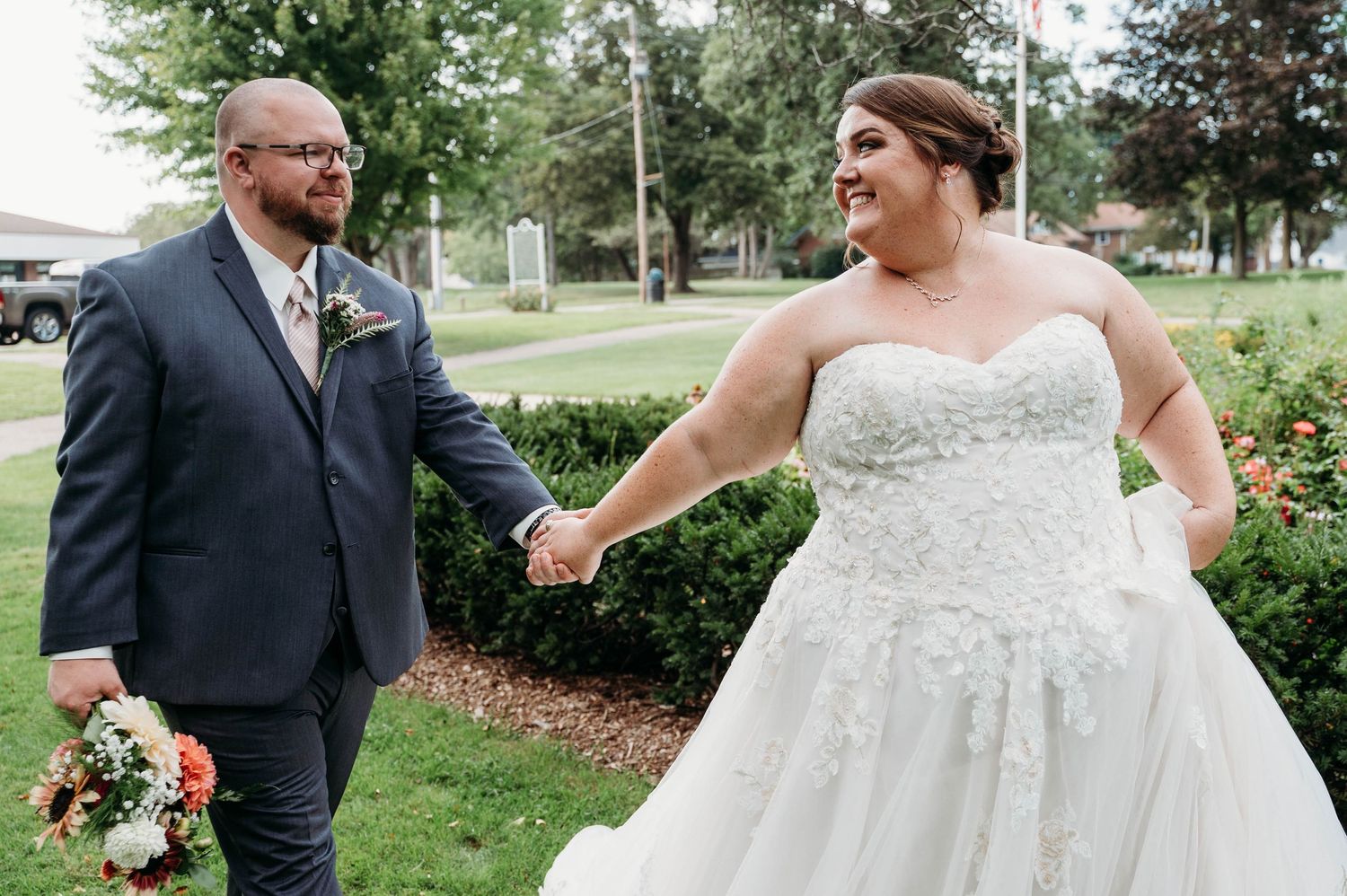 Wedding couple and attendants pose outdoors in a garden setting with green trees and landscaping in background.