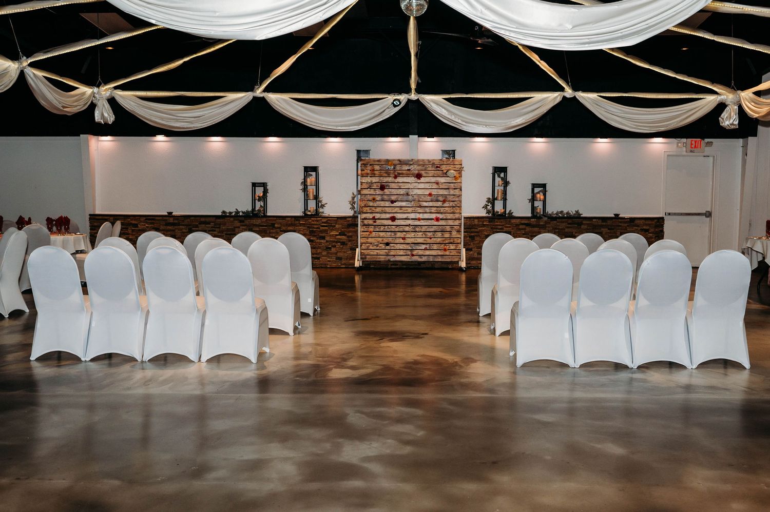 Indoor wedding ceremony setup with white chair covers and draped fabric ceiling in an elegant venue with brick backdrop.