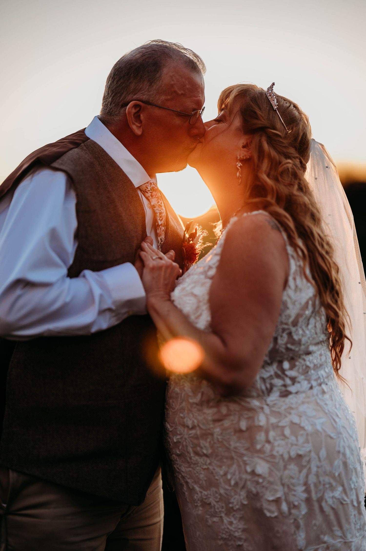 Bride and groom share a romantic sunset kiss, golden backlight glowing behind them at their outdoor wedding ceremony.