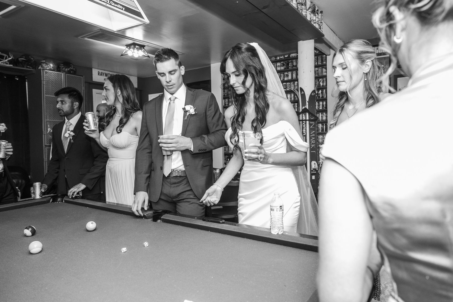Bride and groom playing pool with wedding guests at reception venue bar in black and white photo.